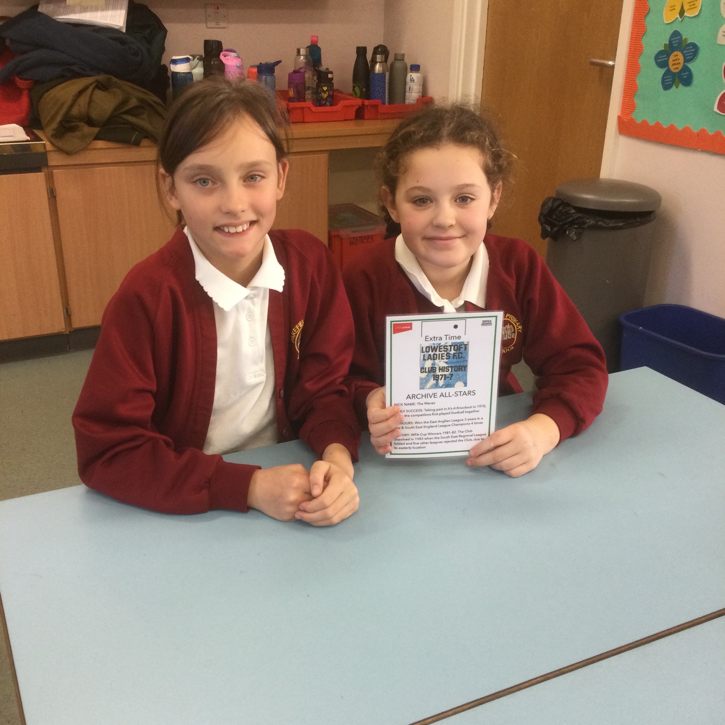 Two school girls face the camera, they are seated at a blue table and holding up a plasticised card about Lowestoft Ladies FC