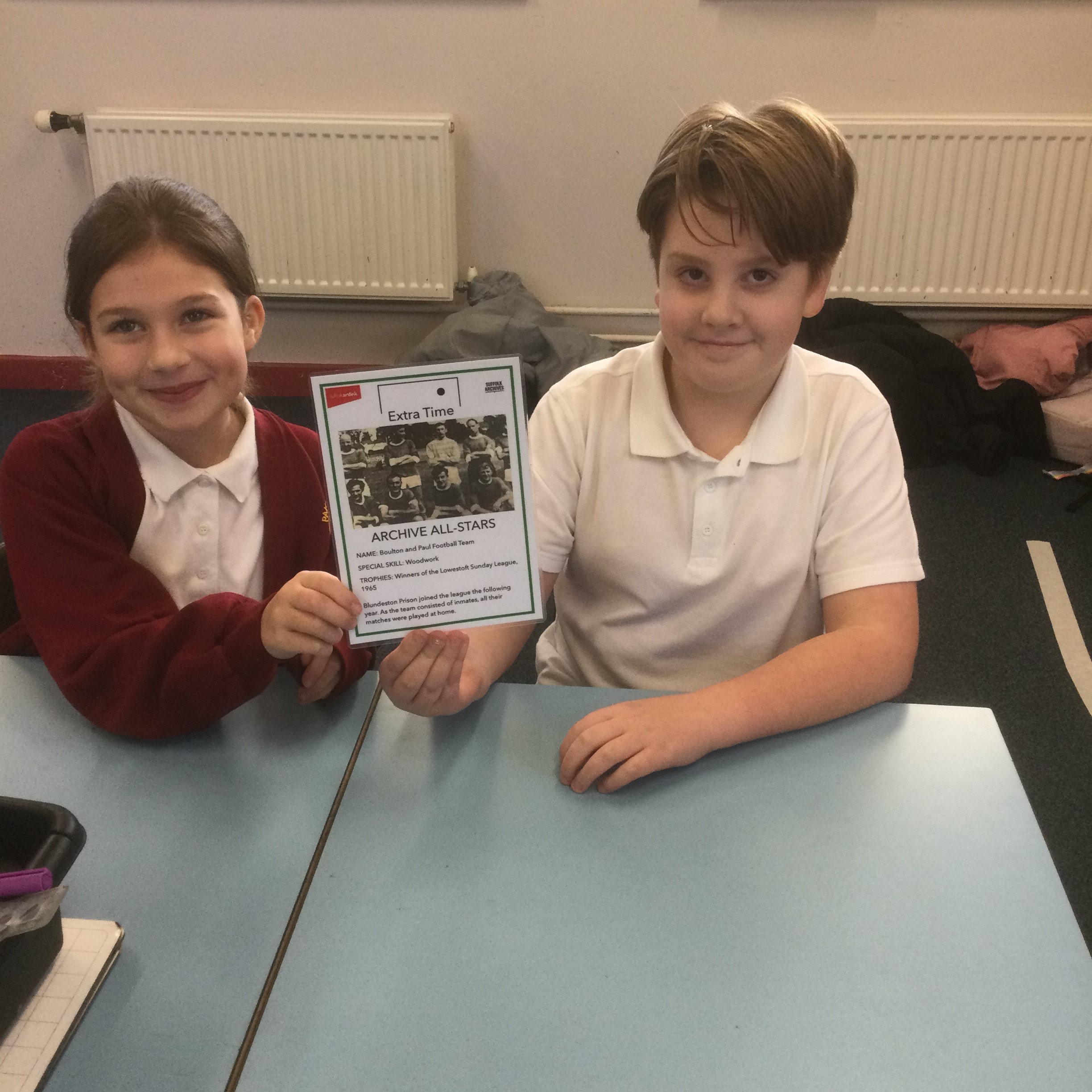 A schoolboy wearing a white T-shirt is seated next to a school girl in a maroon cardigan. They are holding up a card with a monochrome photo of a football team