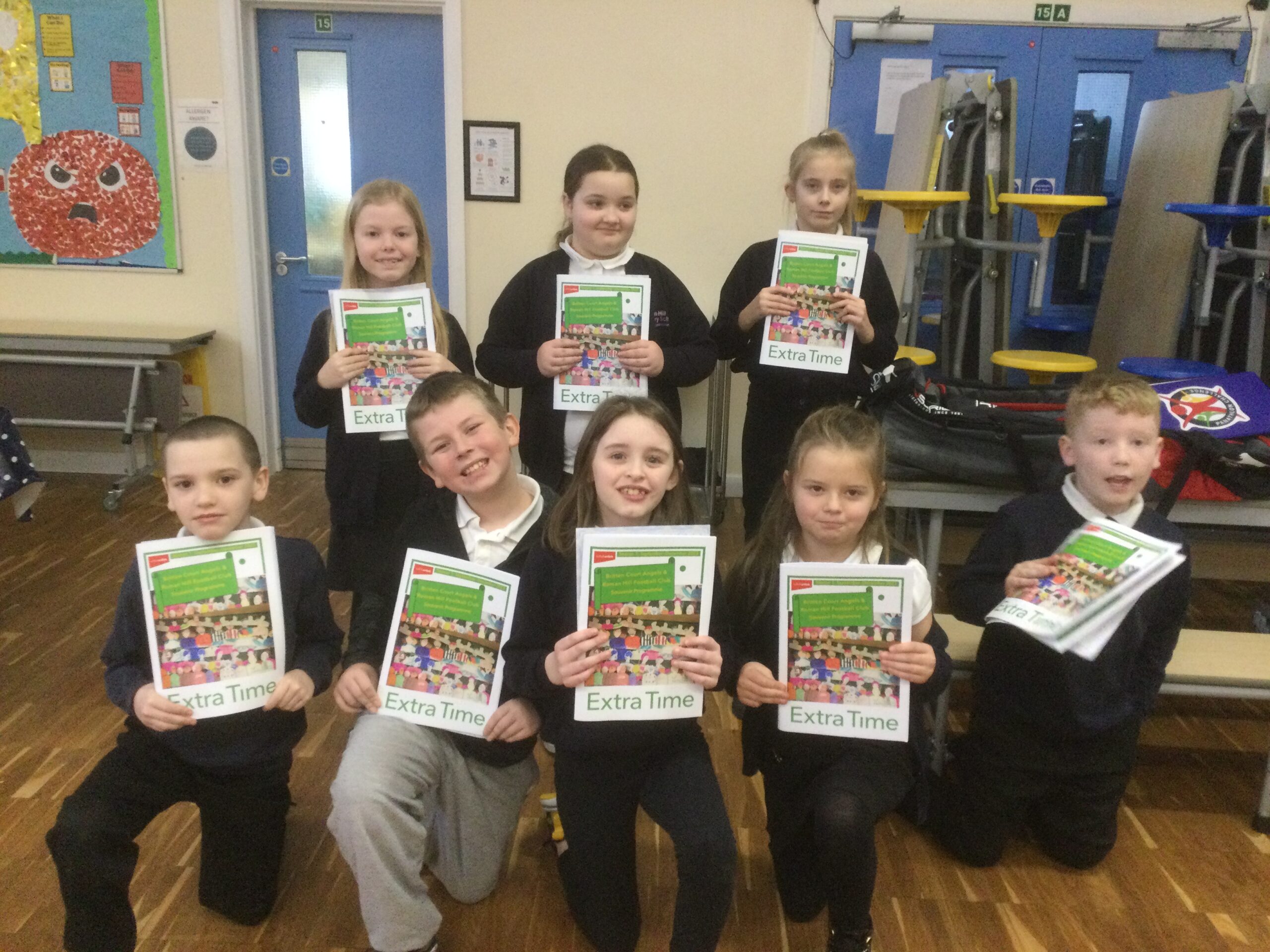 A group of 8 children smiling at the camera and holding their copies of the Extra Time Souvenir Programme