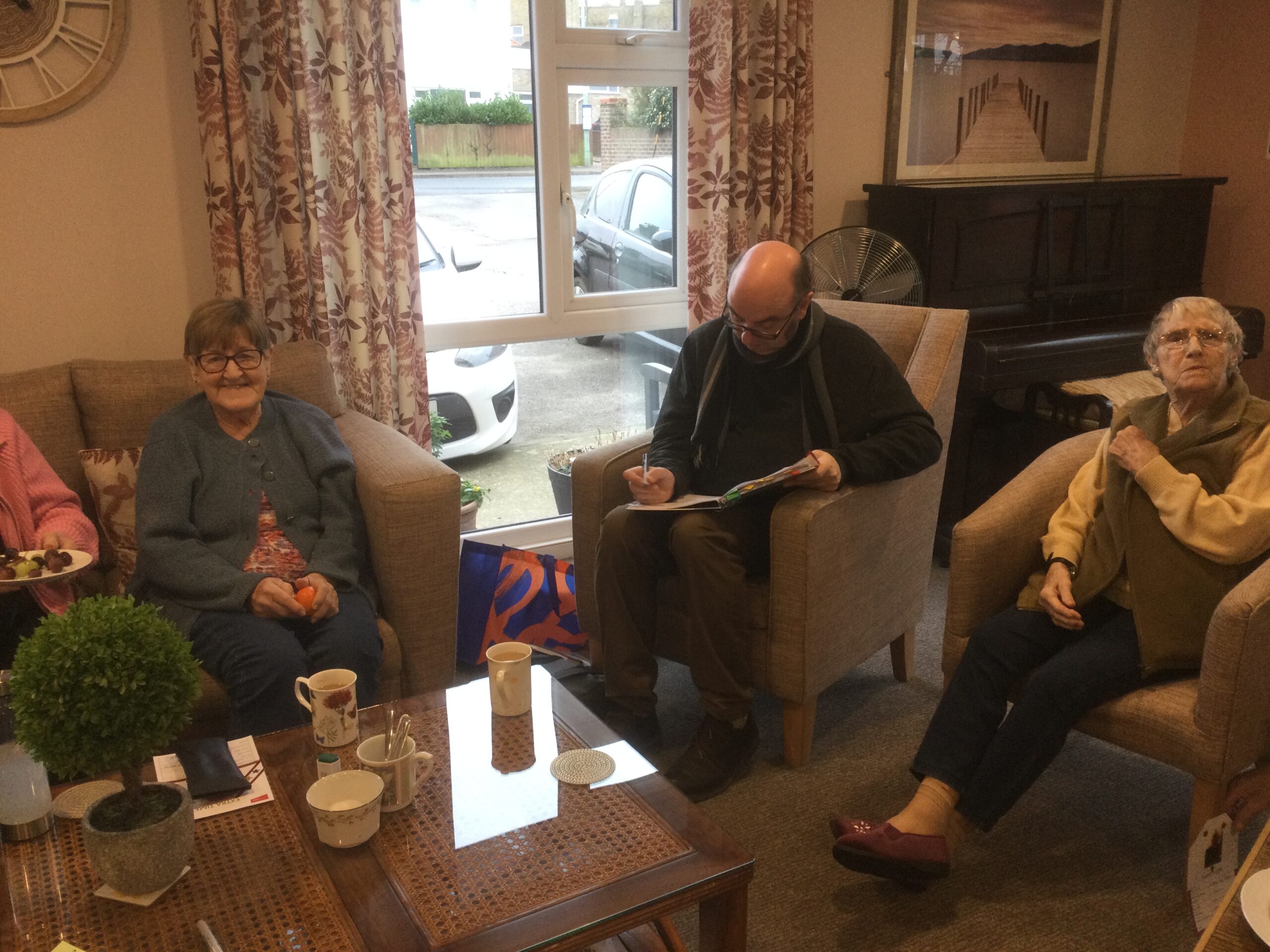 A group of adults seated in armchairs around a coffee table in front of a window. One of the group is looking down at a book in which he is writing and two others smile at the camera