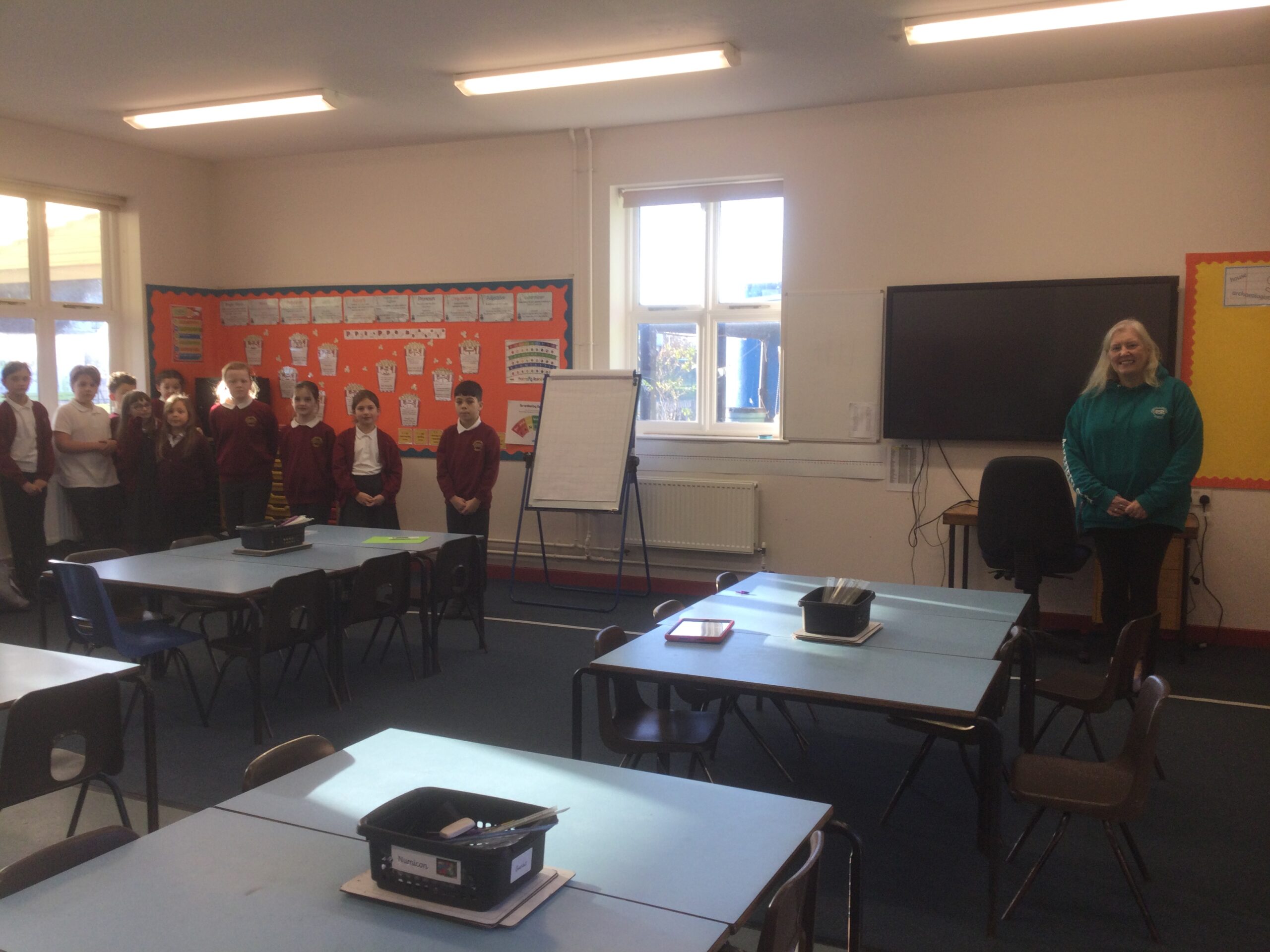 A classroom interior, with desks, flipchart and other learning resources. A length of white tape is laid on the floor and a group of 10 children stand one end, whilst the staff stands at the other end