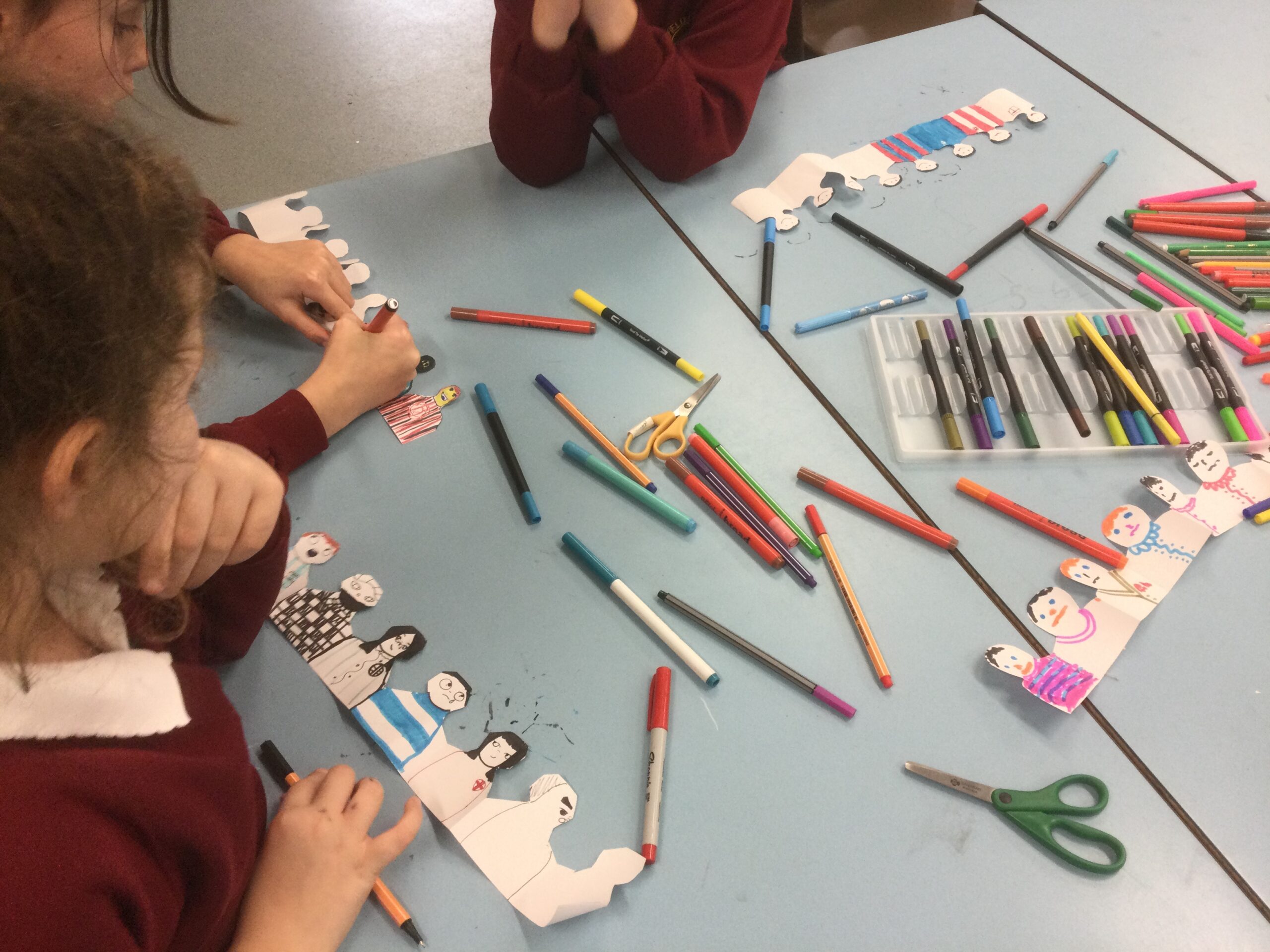 A table covered with coloured pens, with children around it. They are colouring concertina strips of figures to make football spectators