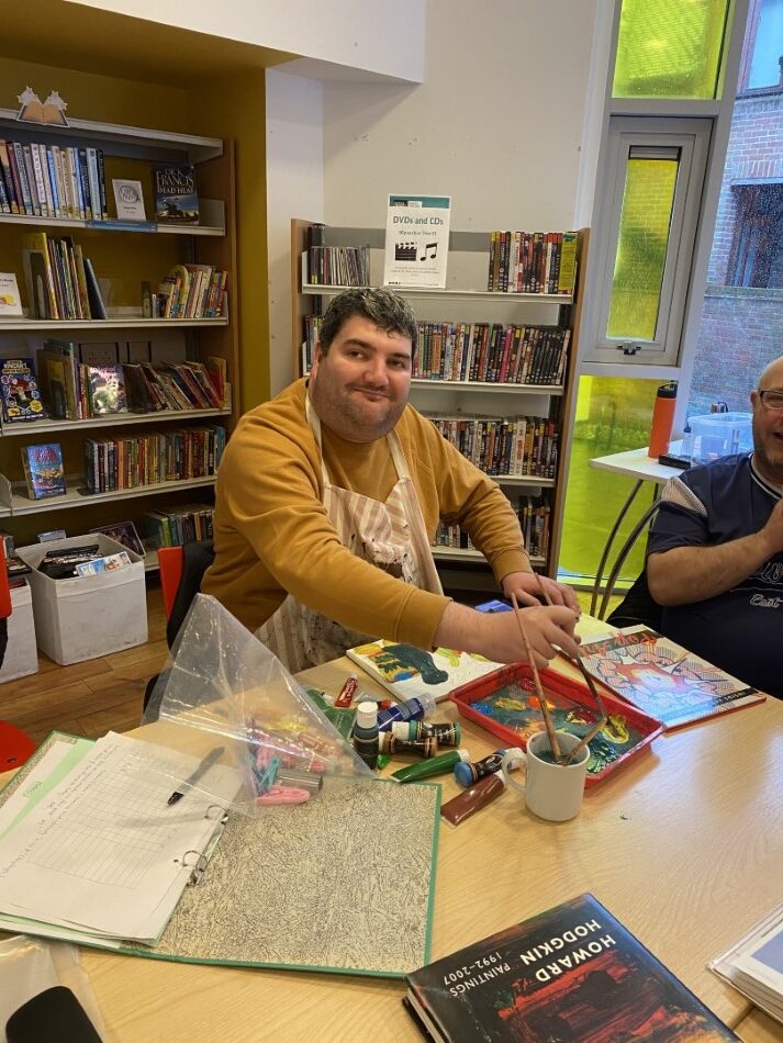 A smiling man wearing a mustard yellow top and an apron is painting,