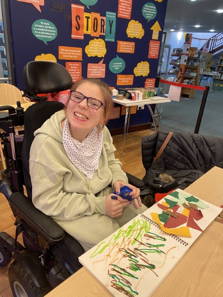 A young woman in a wheelchair is sitting at a table with her sketch book in front of her. On one page is a pen and ink drawing and on the other page is a collage. She is smiling broadly as she looks into the camera.