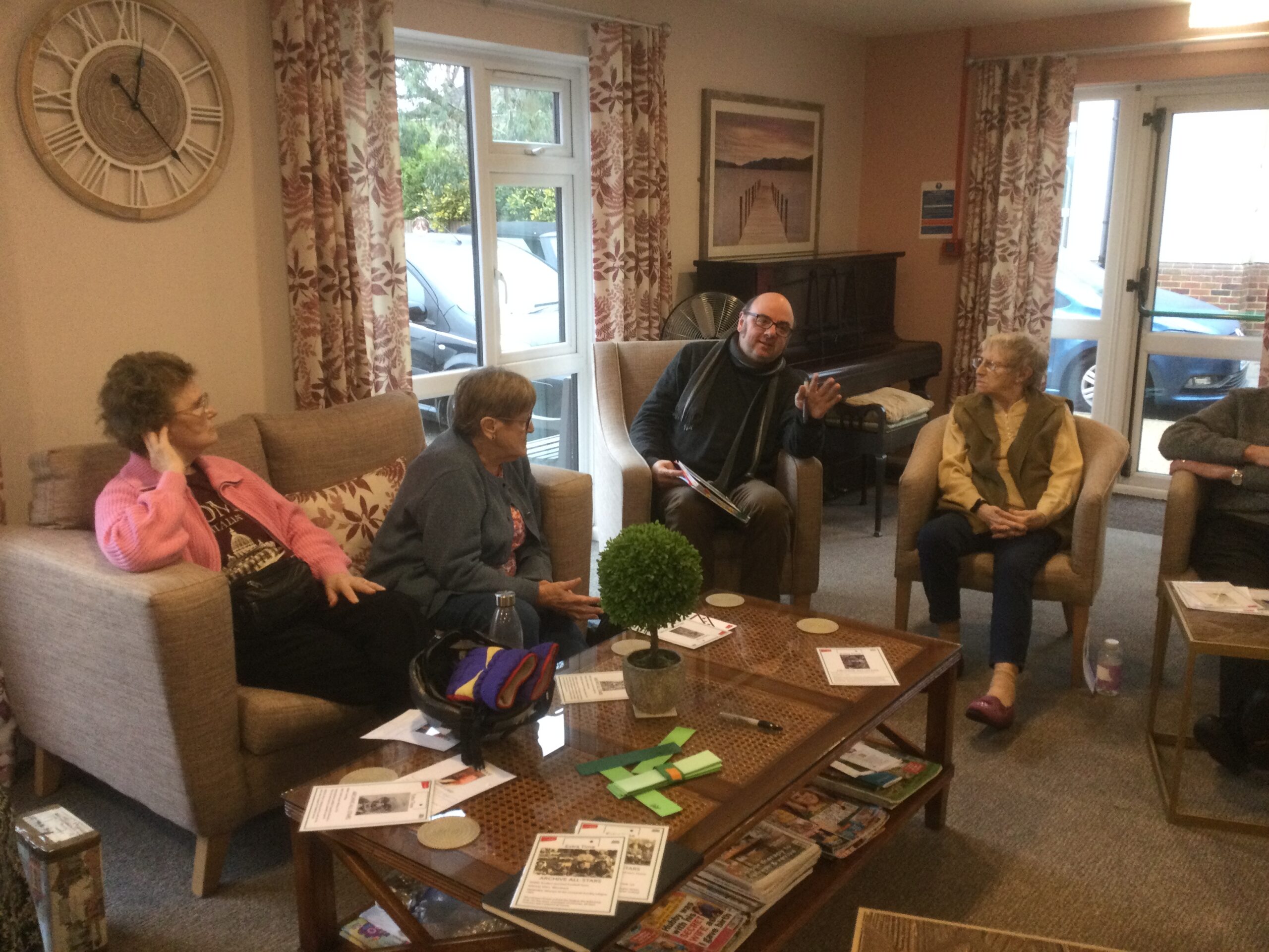 A group of 4 adults seated around a coffee table, 3 or them listening to a man who is leaning slightly in his chair with his arm raised