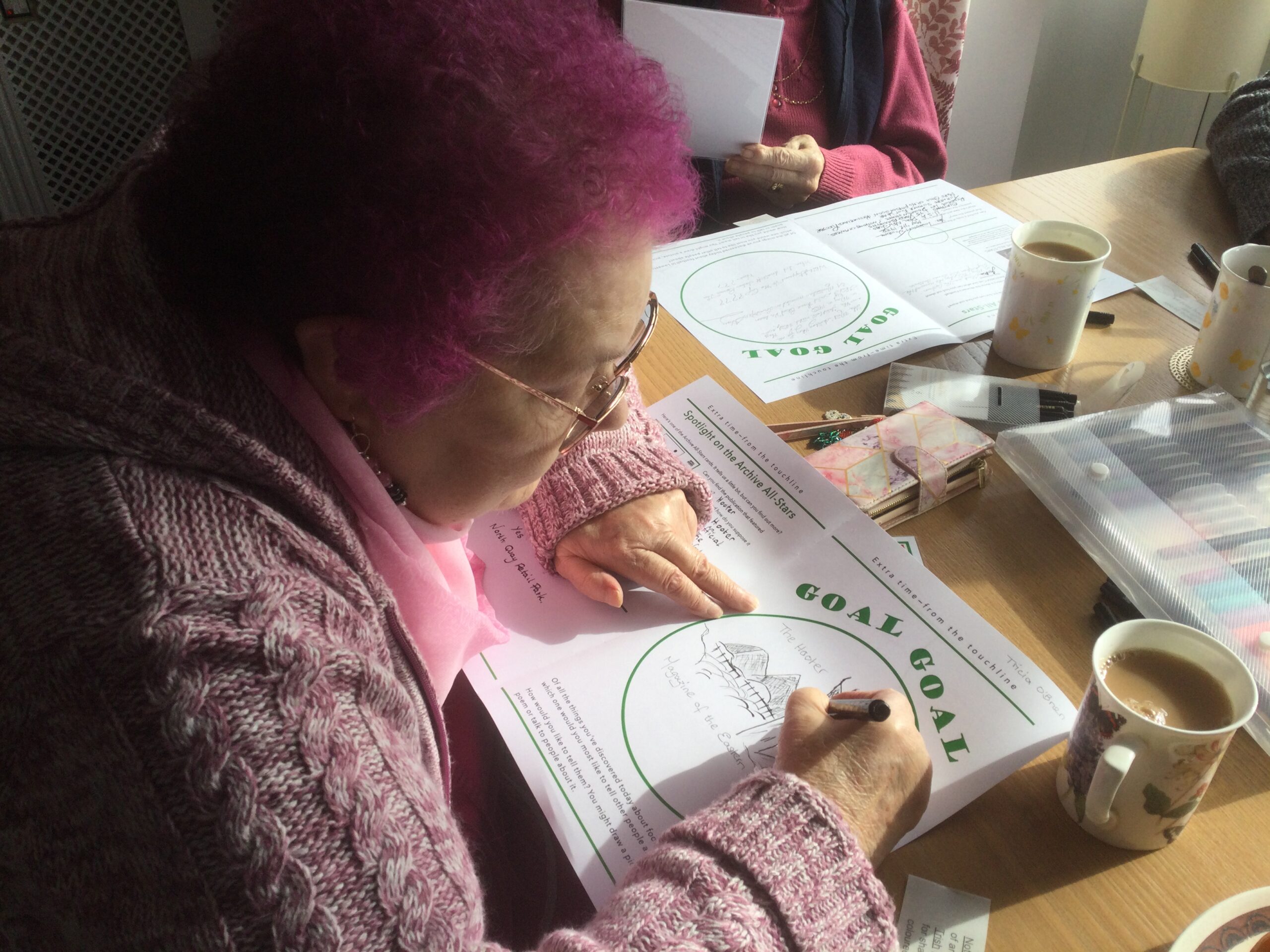 A woman wearing a pink cardigan and scarf and with pink-coloured hair, is drawing an image, surrounded by mugs of tea and coloured pencils