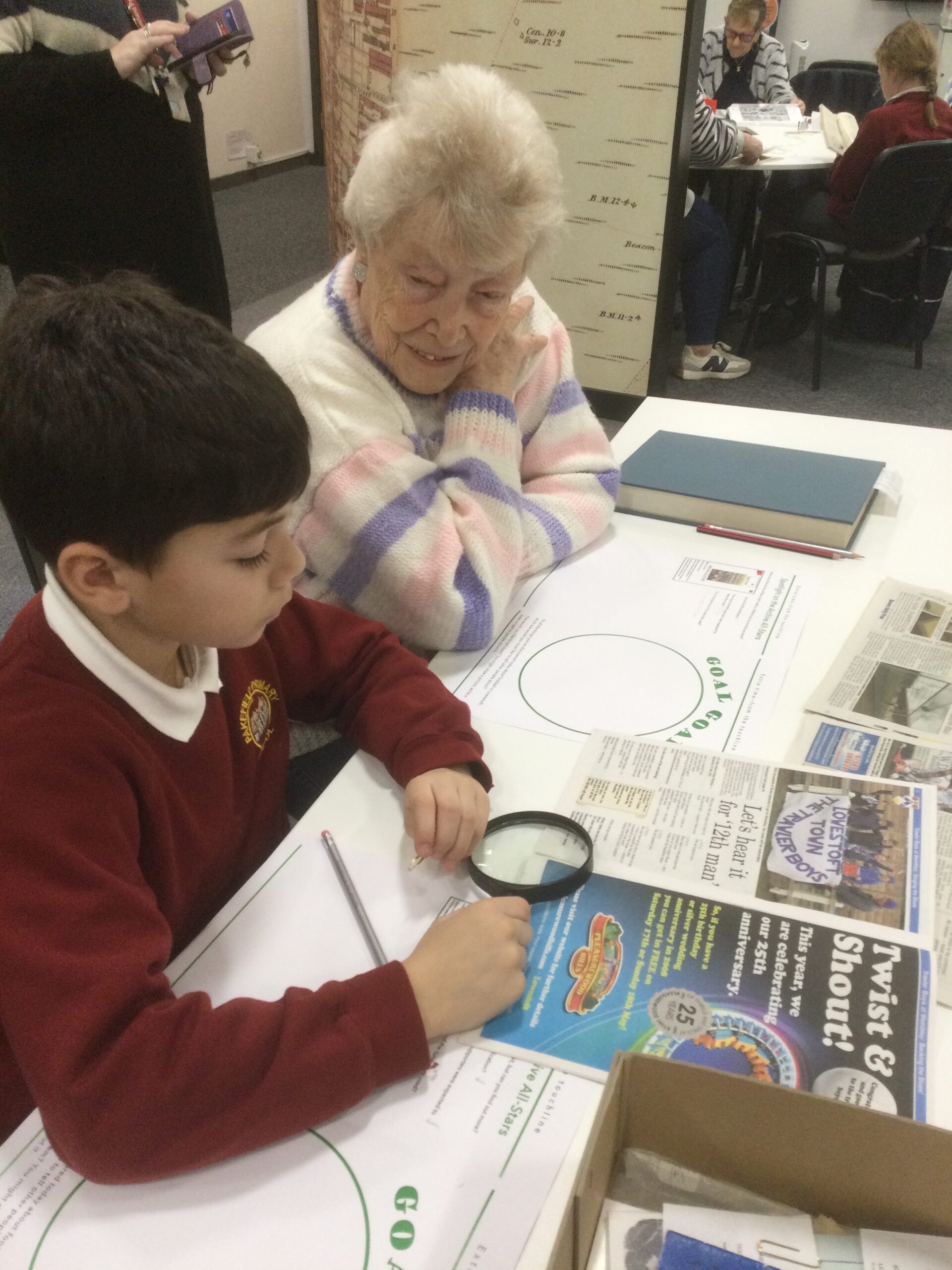 A school boy holding a magnifying glass over a publication with a woman in a striped jumper seated beside him and looking on