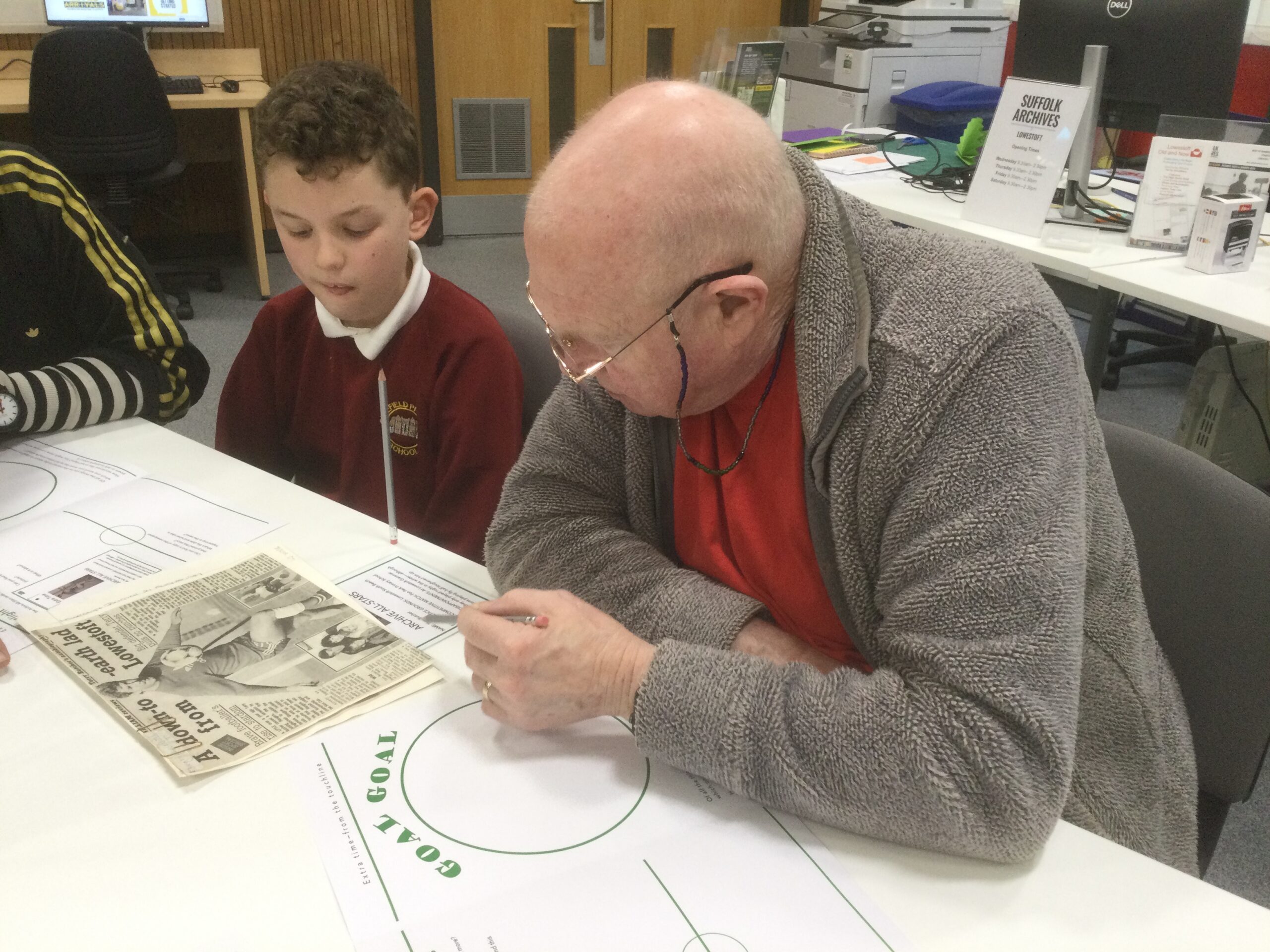 A gentleman and school boy seated at a table, looking at a press cutting of a footballer