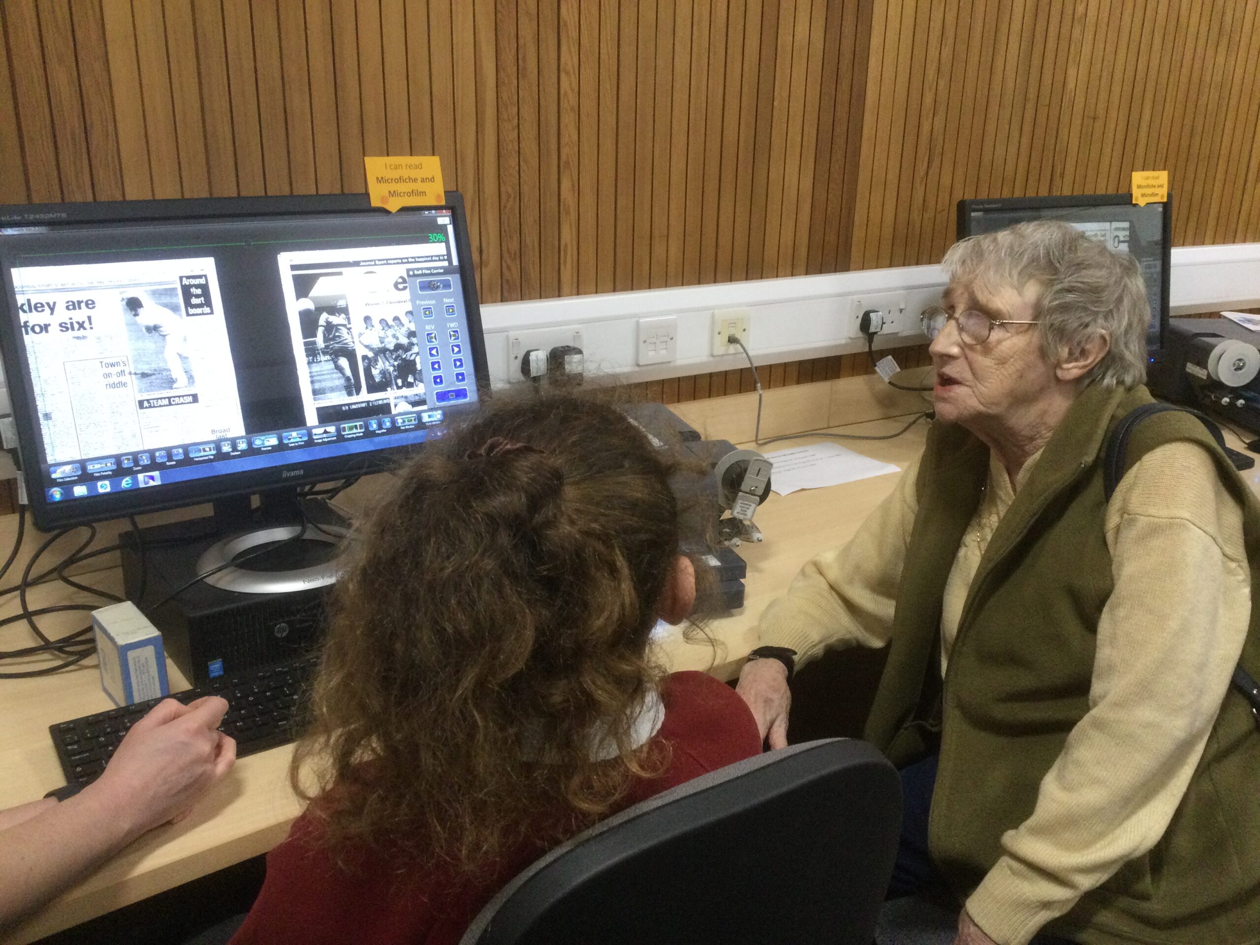 A woman sits beside a school girl in front of a microfiche monitor looking at pictures of footballers.
