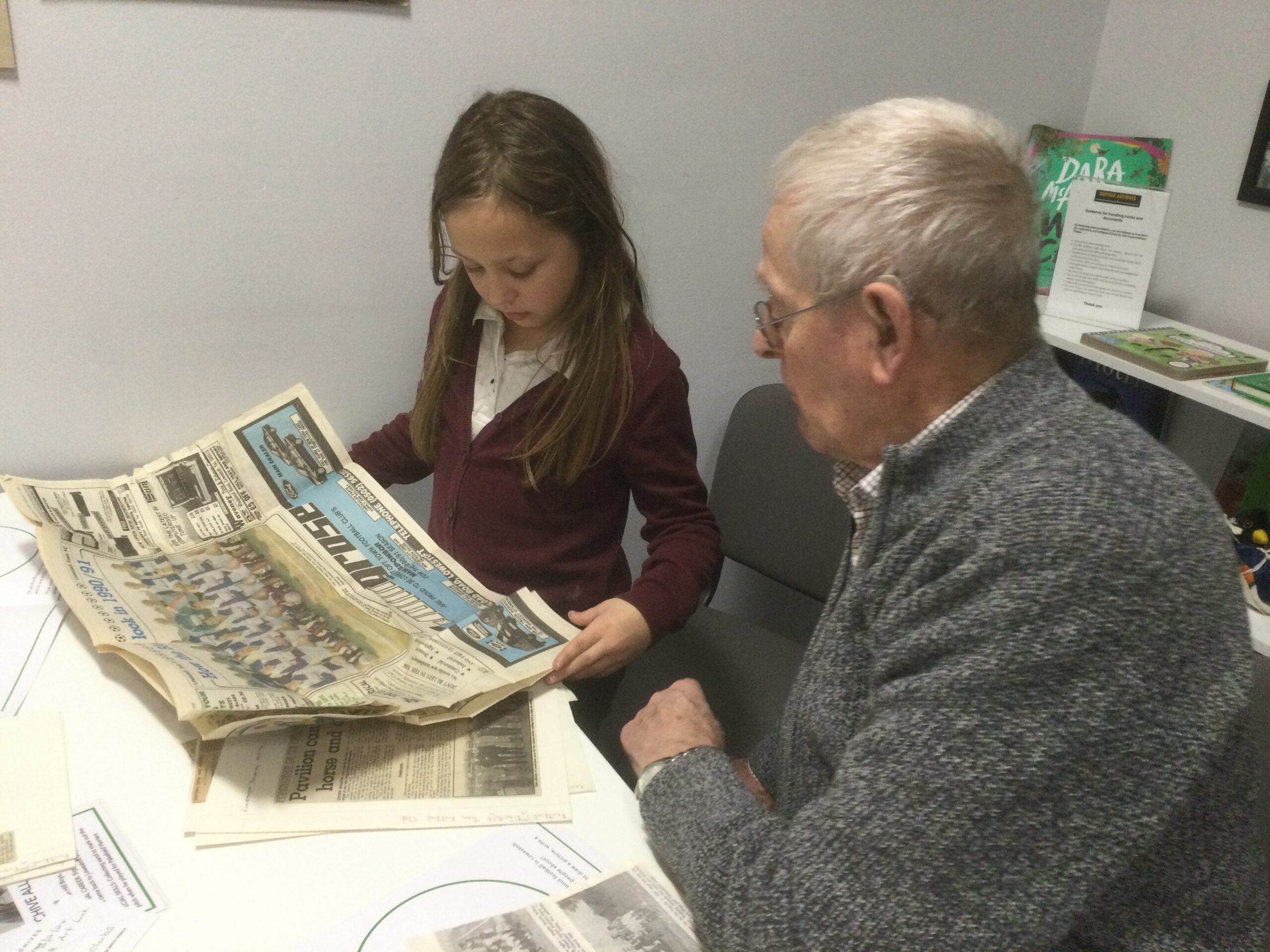 A gentleman seated beside a school girl, both looking at an old newspaper