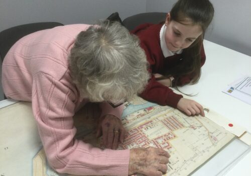 A woman in a pink jumper leans over a map looking closely at an area and pointing something out to the school girl beside her