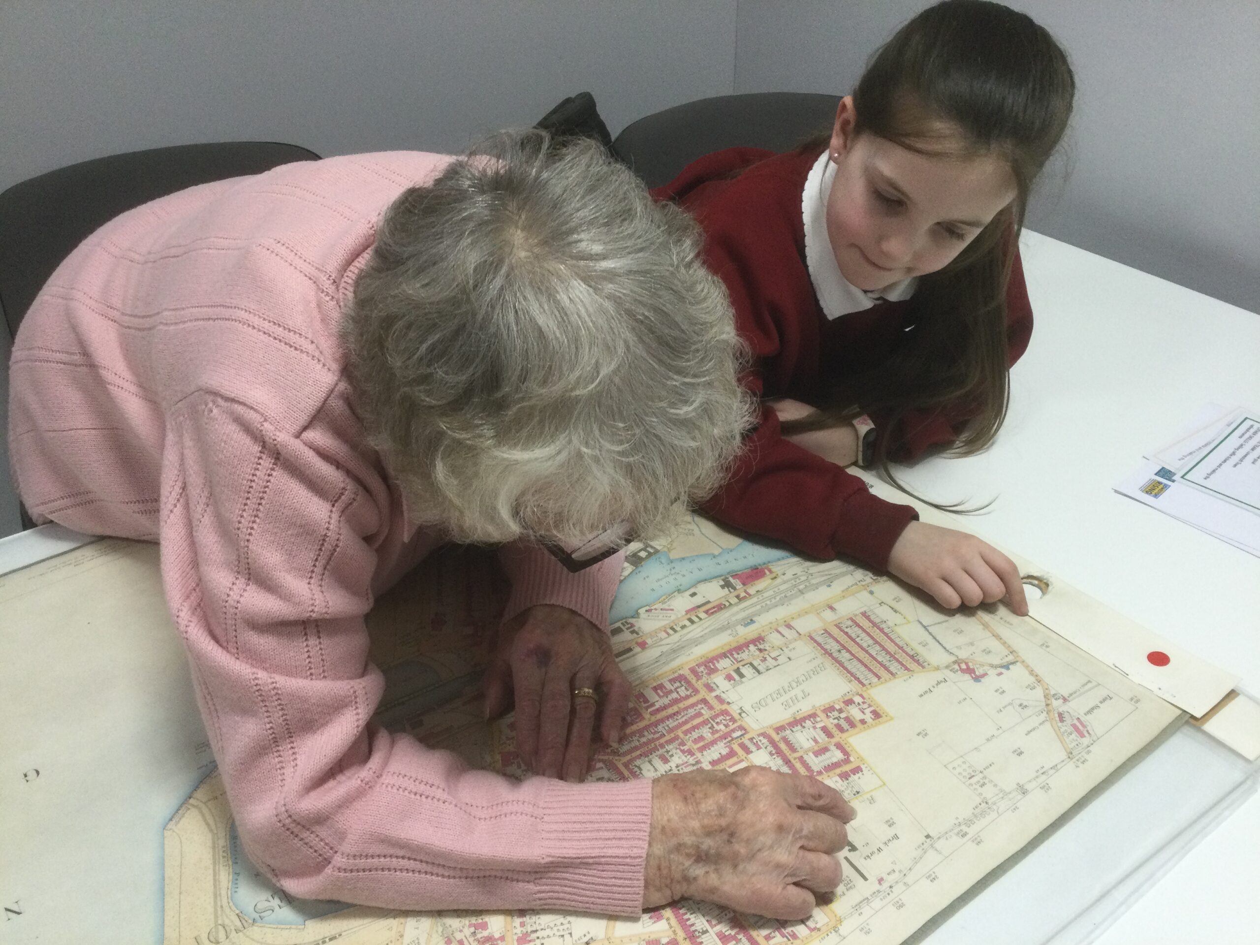 A woman in a pink jumper leans over a map looking closely at an area and pointing something out to the school girl beside her