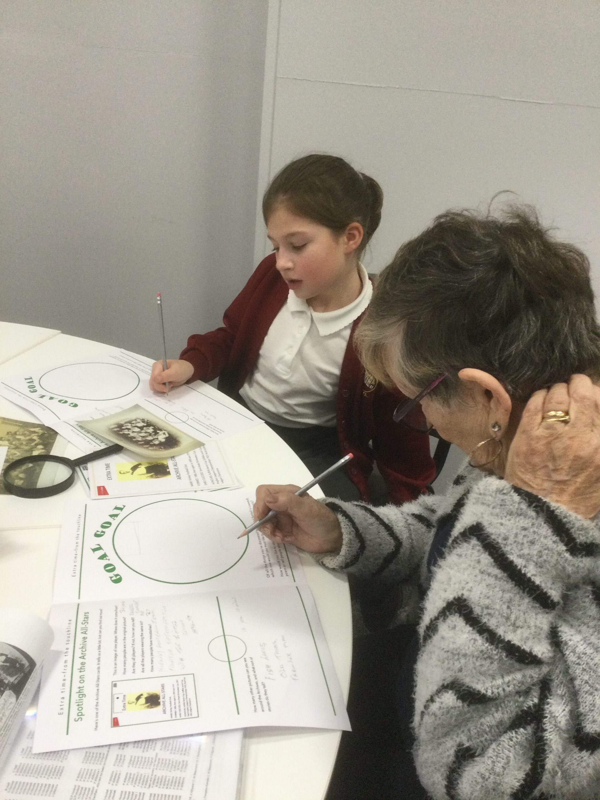 A school girl and adult seated side by side using pencils to note information in their worksheets. An old photograph and magnifying glass is on the table in front of them