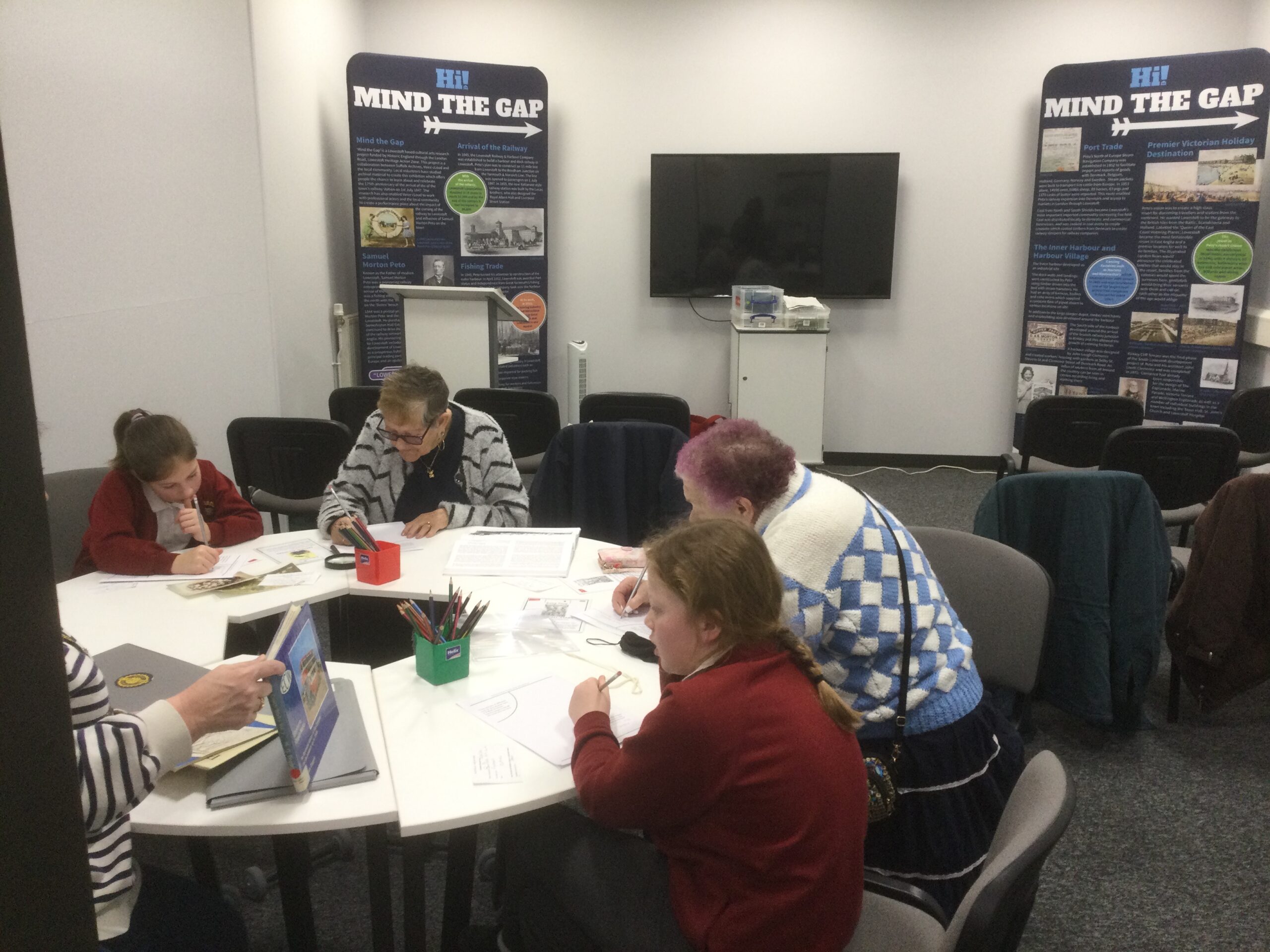 Pairs of adults and children seated at a circular table, a pot of coloured pencils in front of them, writing down notes