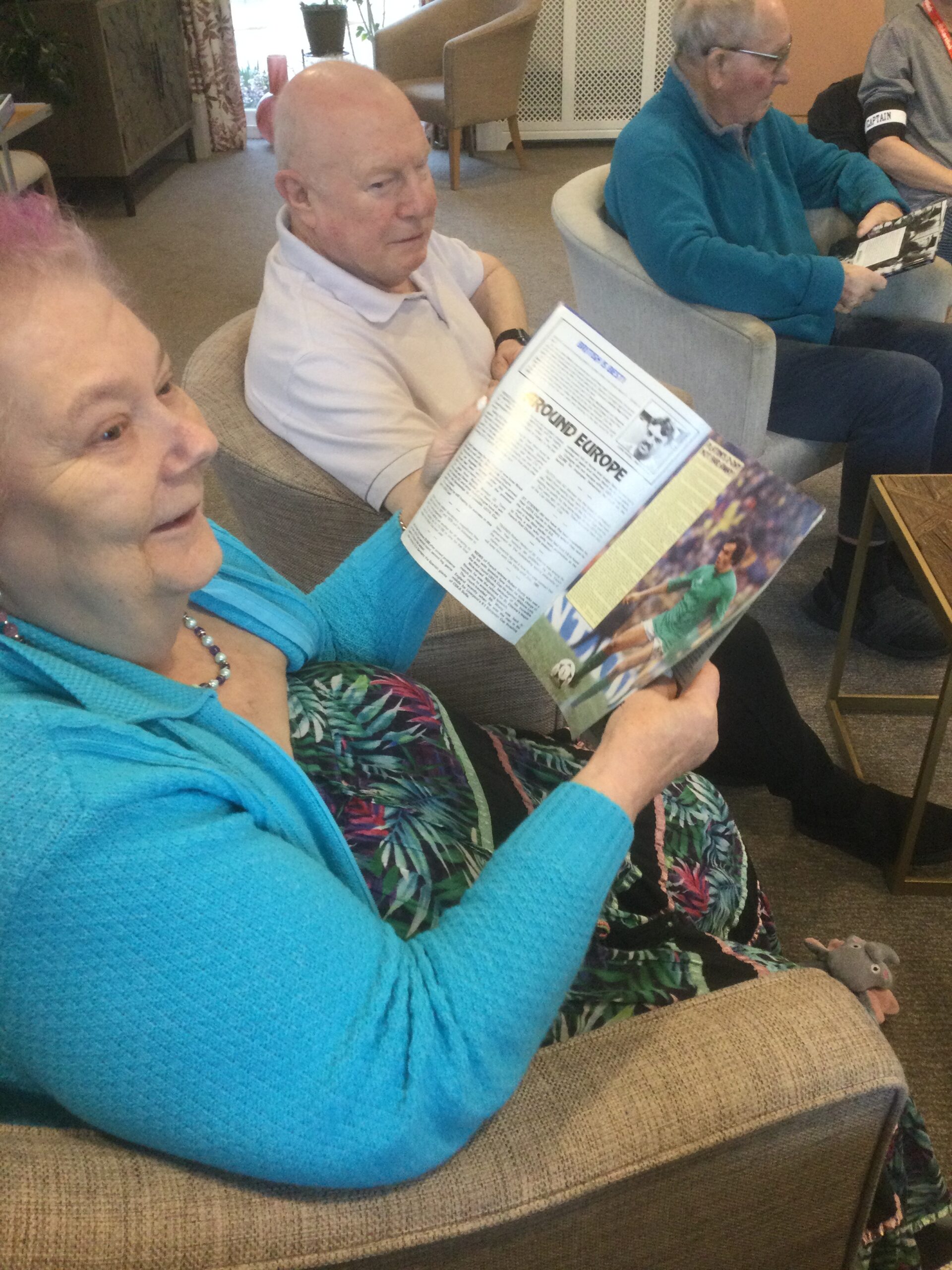 Woman seated in armchair, holding footballing programme open with text and photo of player. Two men seated in armchairs to her left