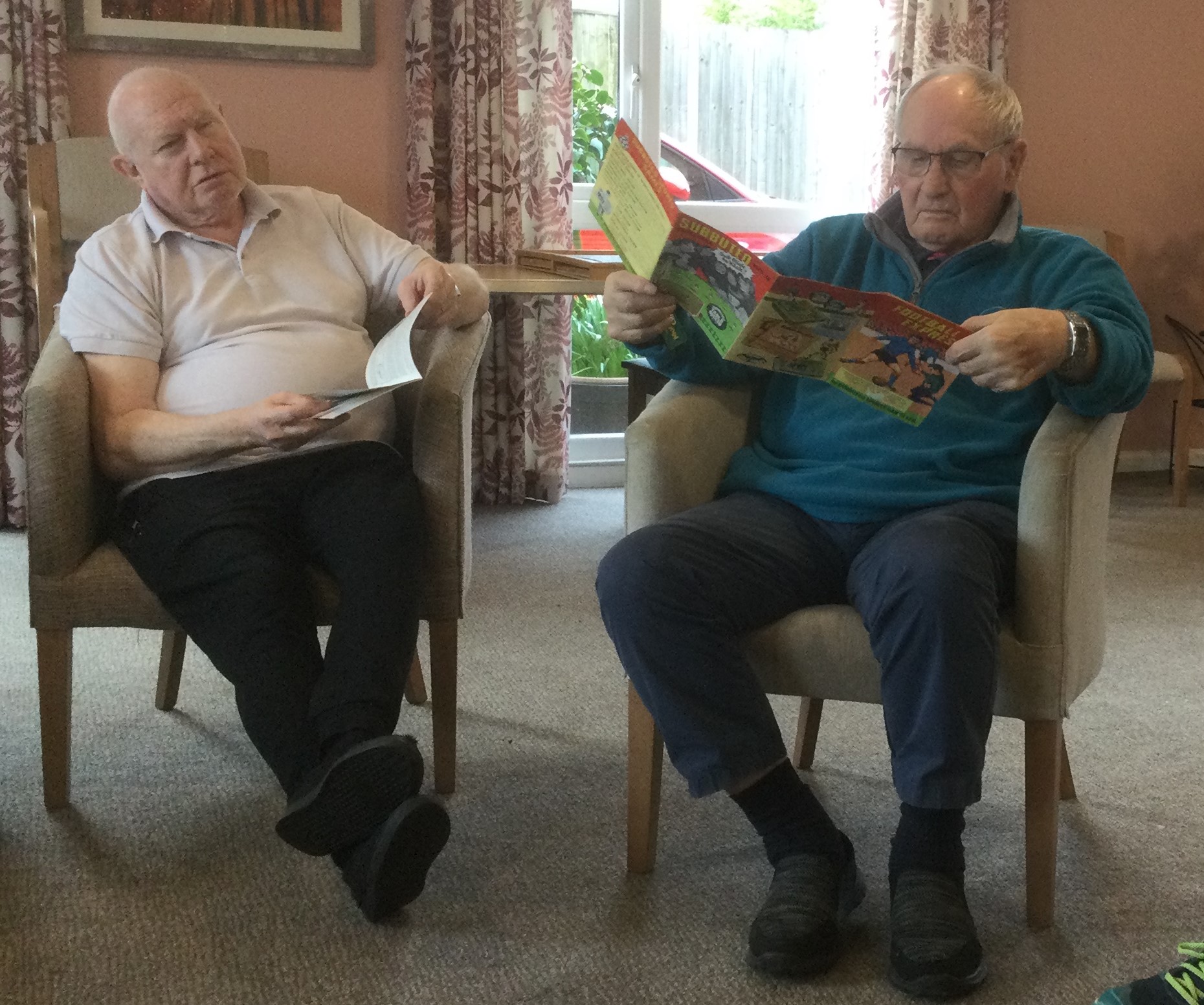 Two gentlemen seated in easy chairs reading football programmes