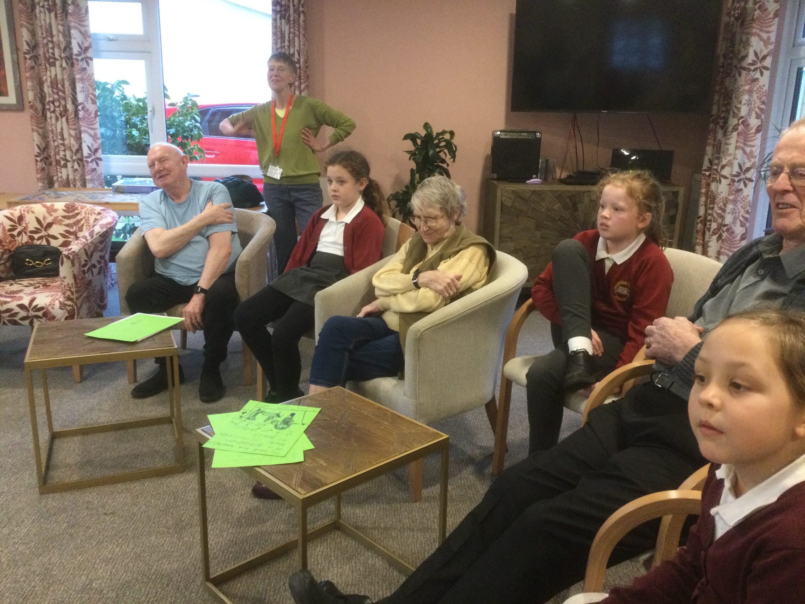 A mixed group of adults and school children, seated in a semi-circle all focused on something out of camera shot