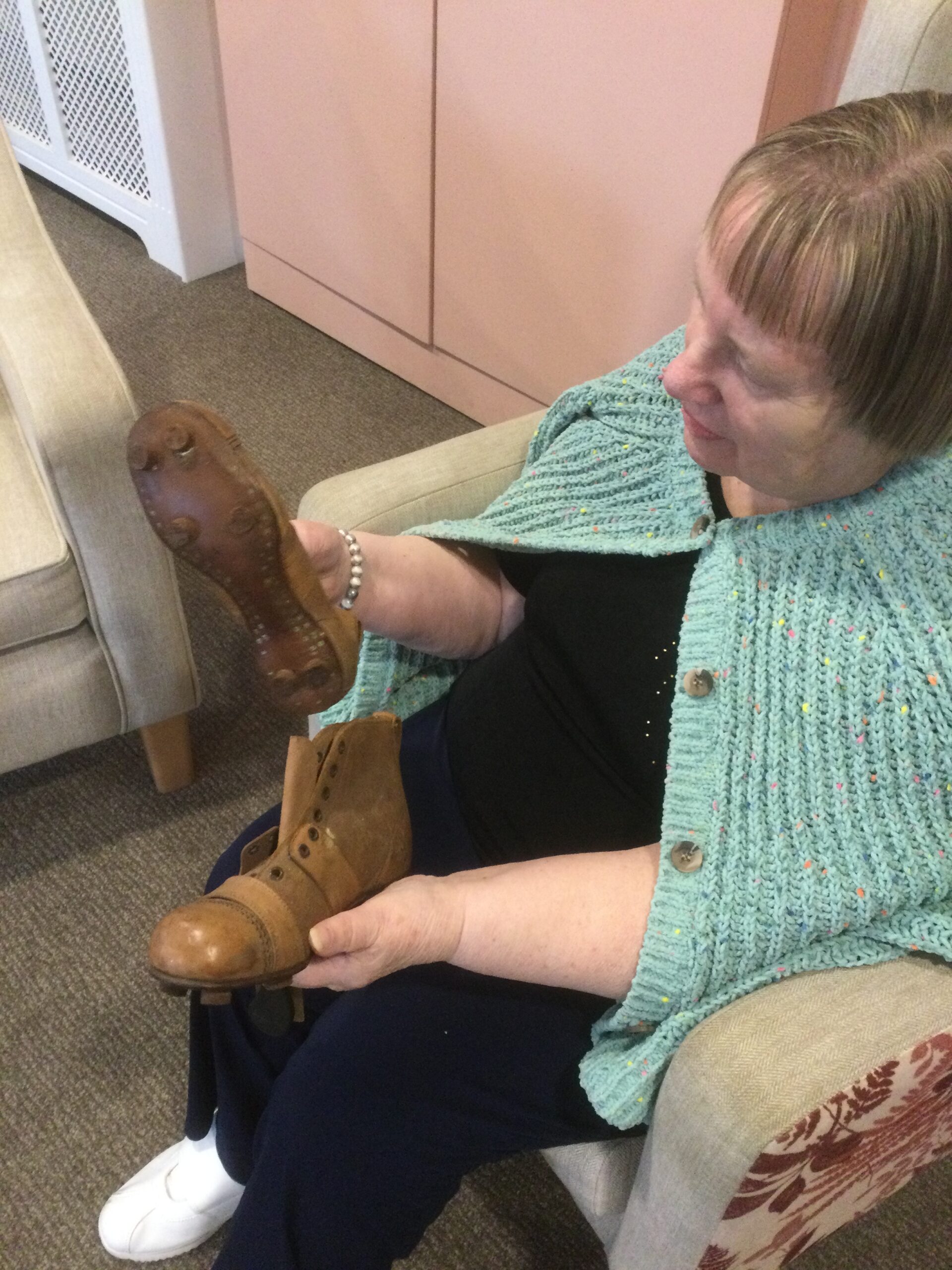 A woman seated, wearing a light green cardigan over her shoulders and holding a pair of old leather football boots