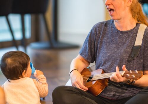 Abby Page playing a ukelele watched by a small child.