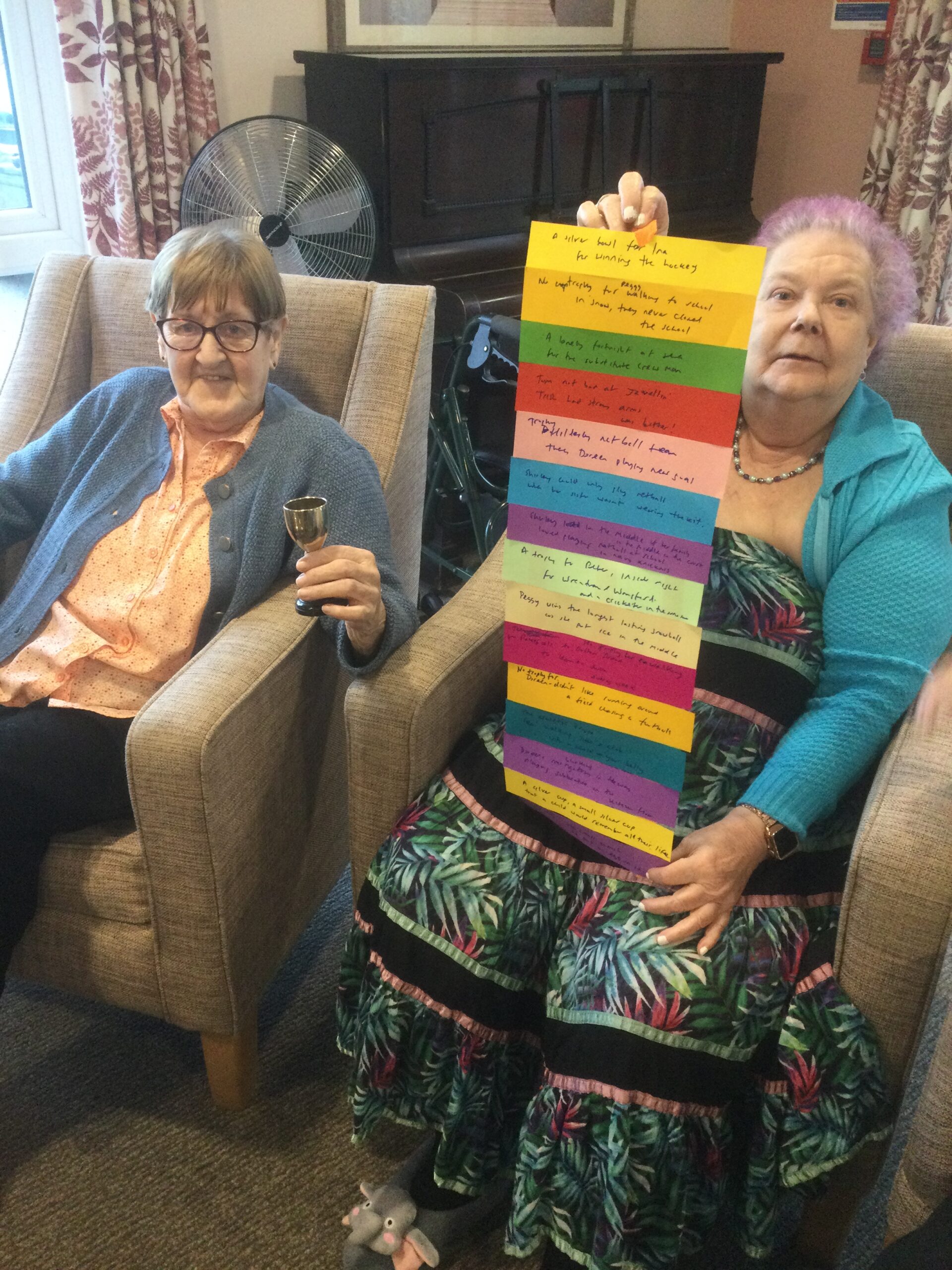 Two women seated side by side, one holding a small silver cup in her hand whilst the other holds up a long strip of different coloured paper strips, with hand written lines on them