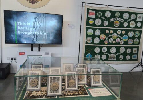 A large screen on the wall shows a female footballer and the text Bringing Heritage to Life. Beside is suspended a large football banner covered with colourful circular images and text. In the forefront is a clear cabinet with framed photographs and clay plaques displayed