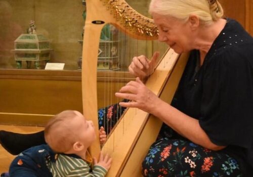 Xenia Horne playing harp on the floor while a baby watches.