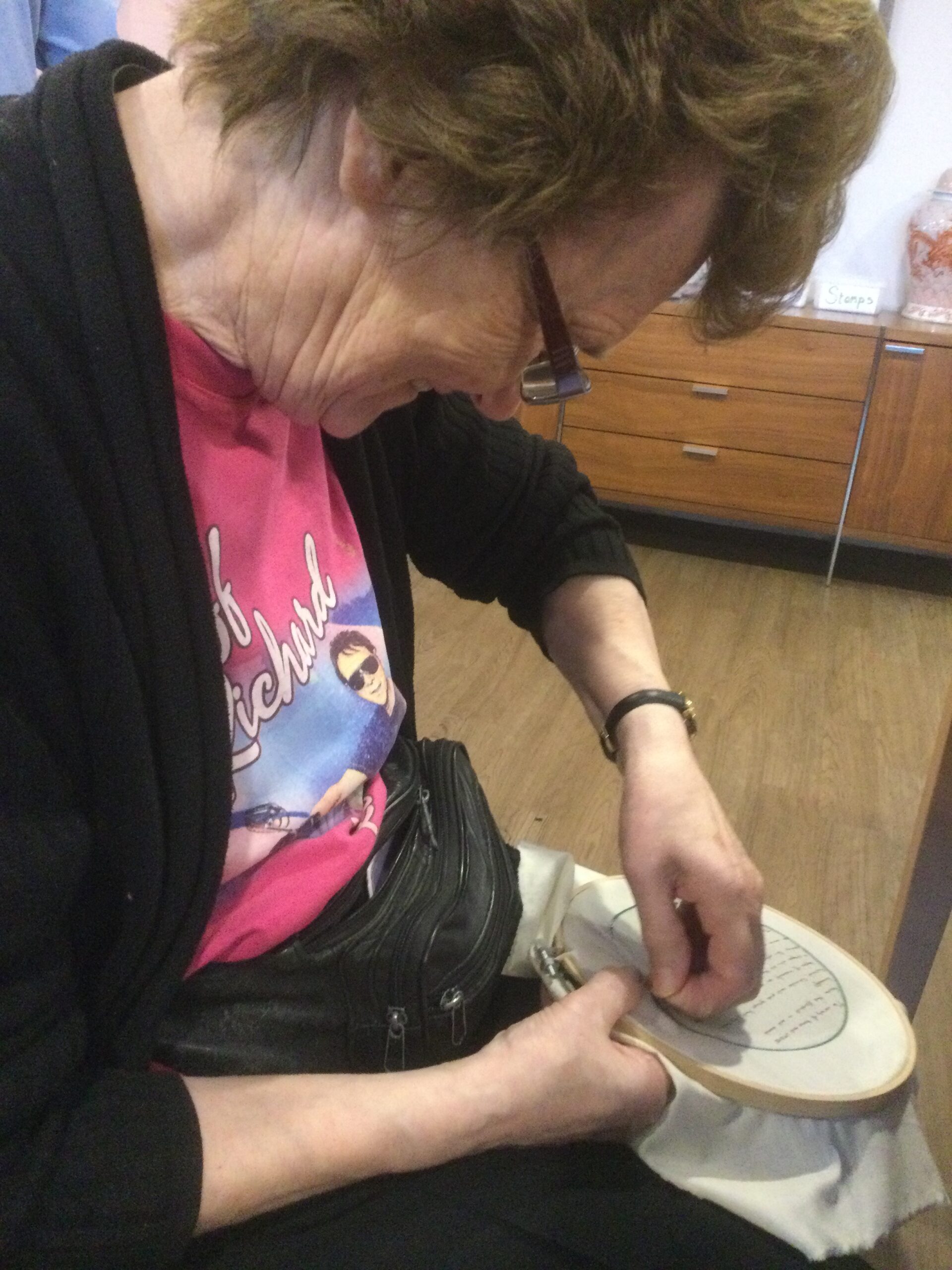 A woman wearing a black cardigan sits, stitching a piece of embroidery