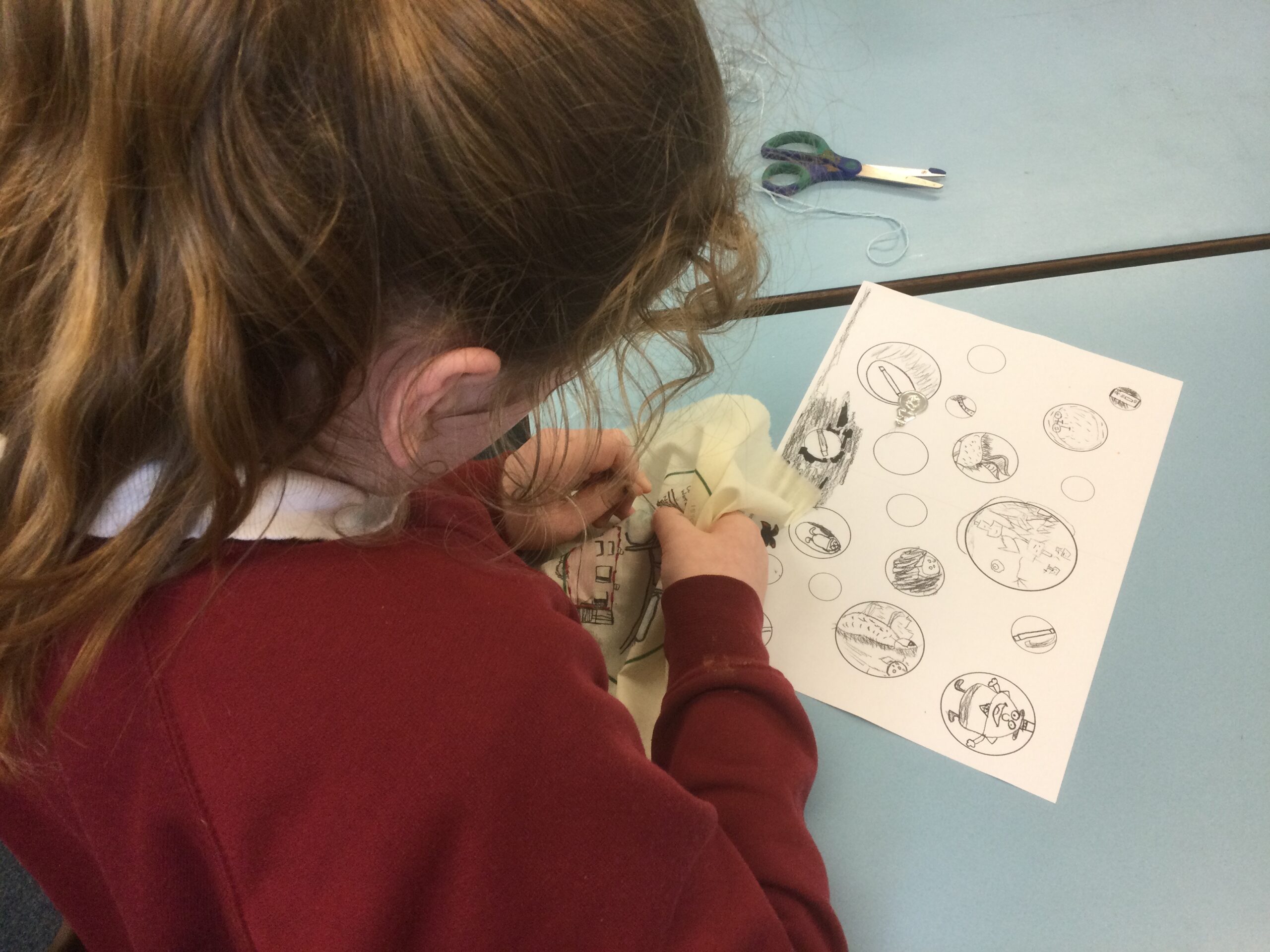 A girl hunched over her embroidery and on the table in front of her is a copy of her bubble drawings