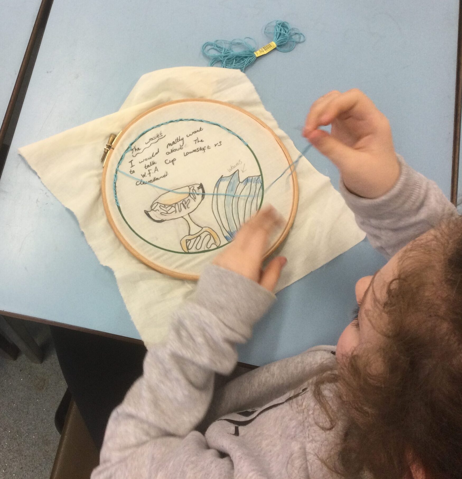 Overhead view of school girl stitching with blue thread on a printed picture of a trophy and a blue wave