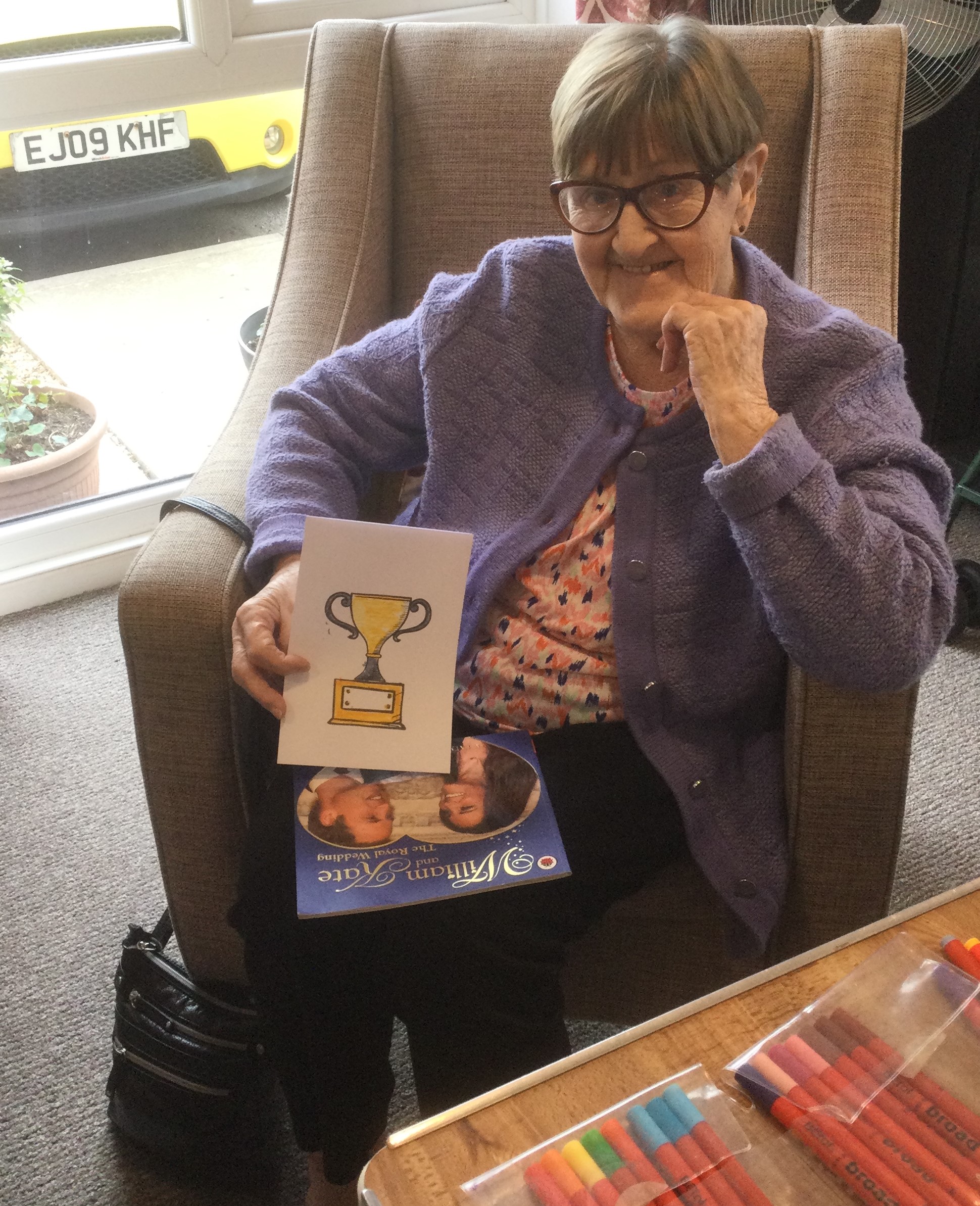 A woman seated in an armchair, holding a card, with rows of coloured pens on the table in front of her