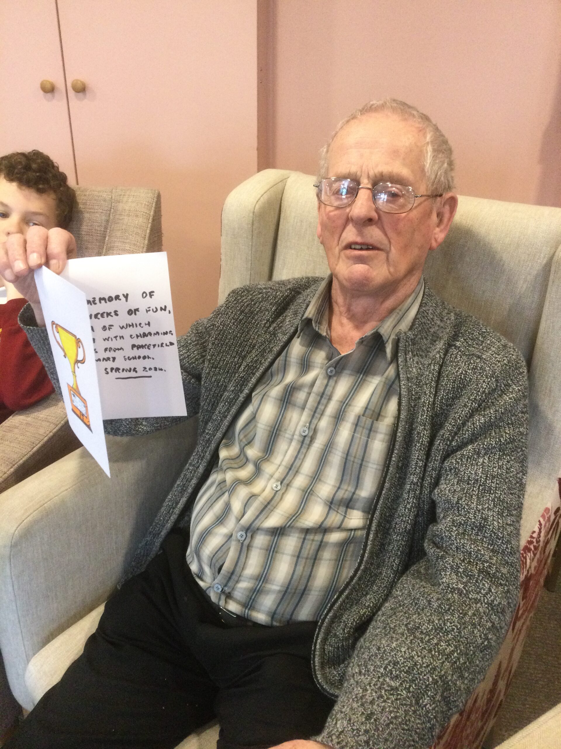 A gentleman seated in an armchair, holding up a hand written card