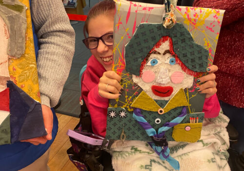 A smiling young woman in a wheel chair holds up her collage self portrait. She is peering around the artwork and smiling.