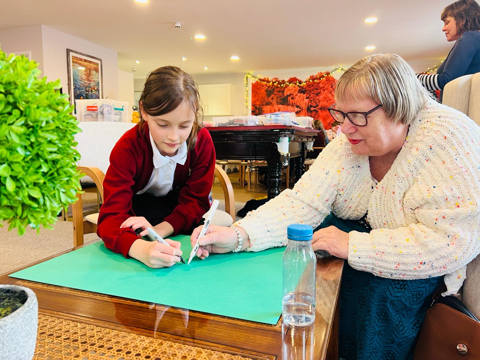 A school child and adult leaning over a large piece of green paper one drawing with a black pen and the other with a white pen