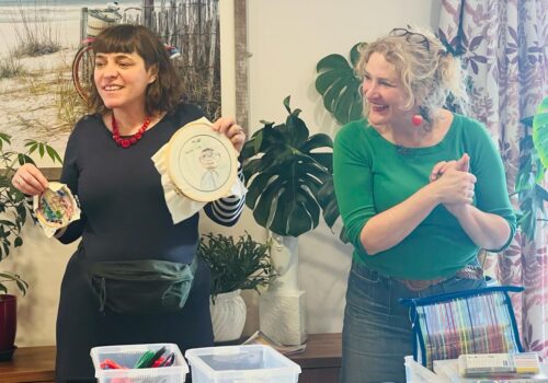 A woman dressed in dark clothes and wearing a large red bead necklace stands next to a woman with light hair and wearing a green top. They are holding up some embroidery and smiling broadly