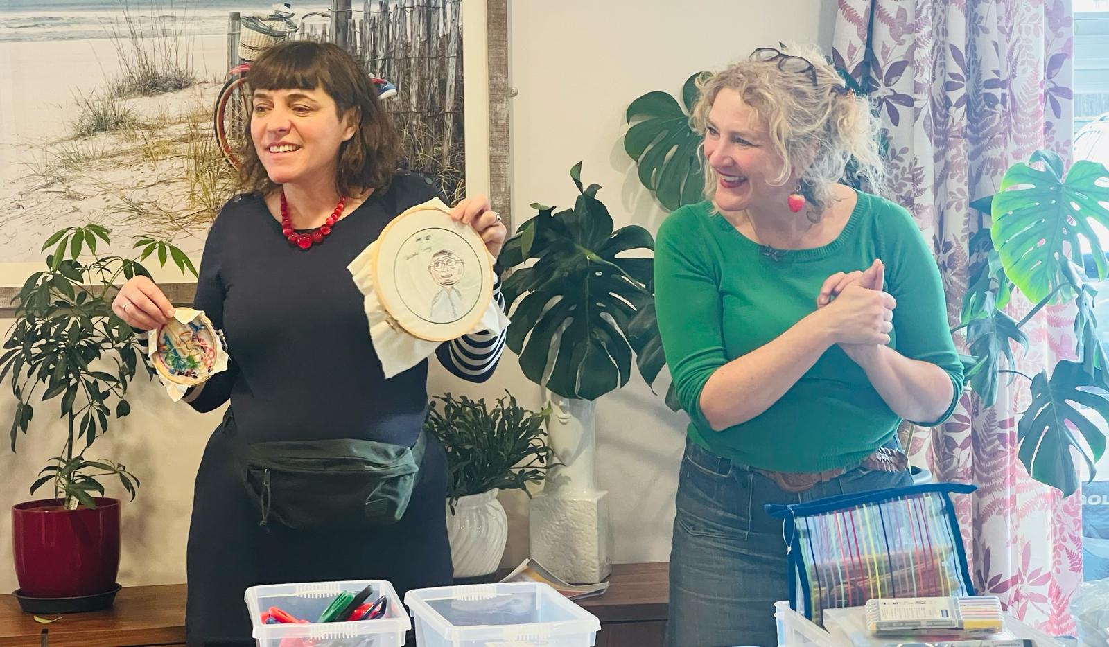 A woman dressed in dark clothes and wearing a large red bead necklace stands next to a woman with light hair and wearing a green top. They are holding up some embroidery and smiling broadly