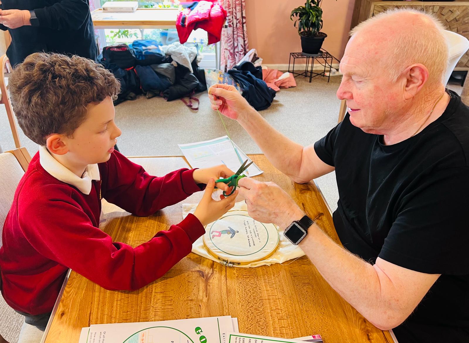 A gentleman sits on right holding a length of green cotton thread between his fingers while a school boy opposite uses a pair of green handled scissors to cut the thread