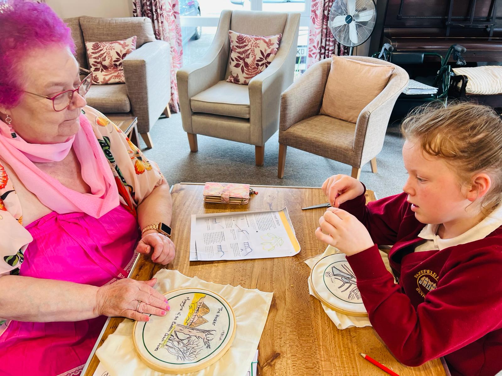 An adult female dressed predominantly in pink sits opposite a school child threading a needle