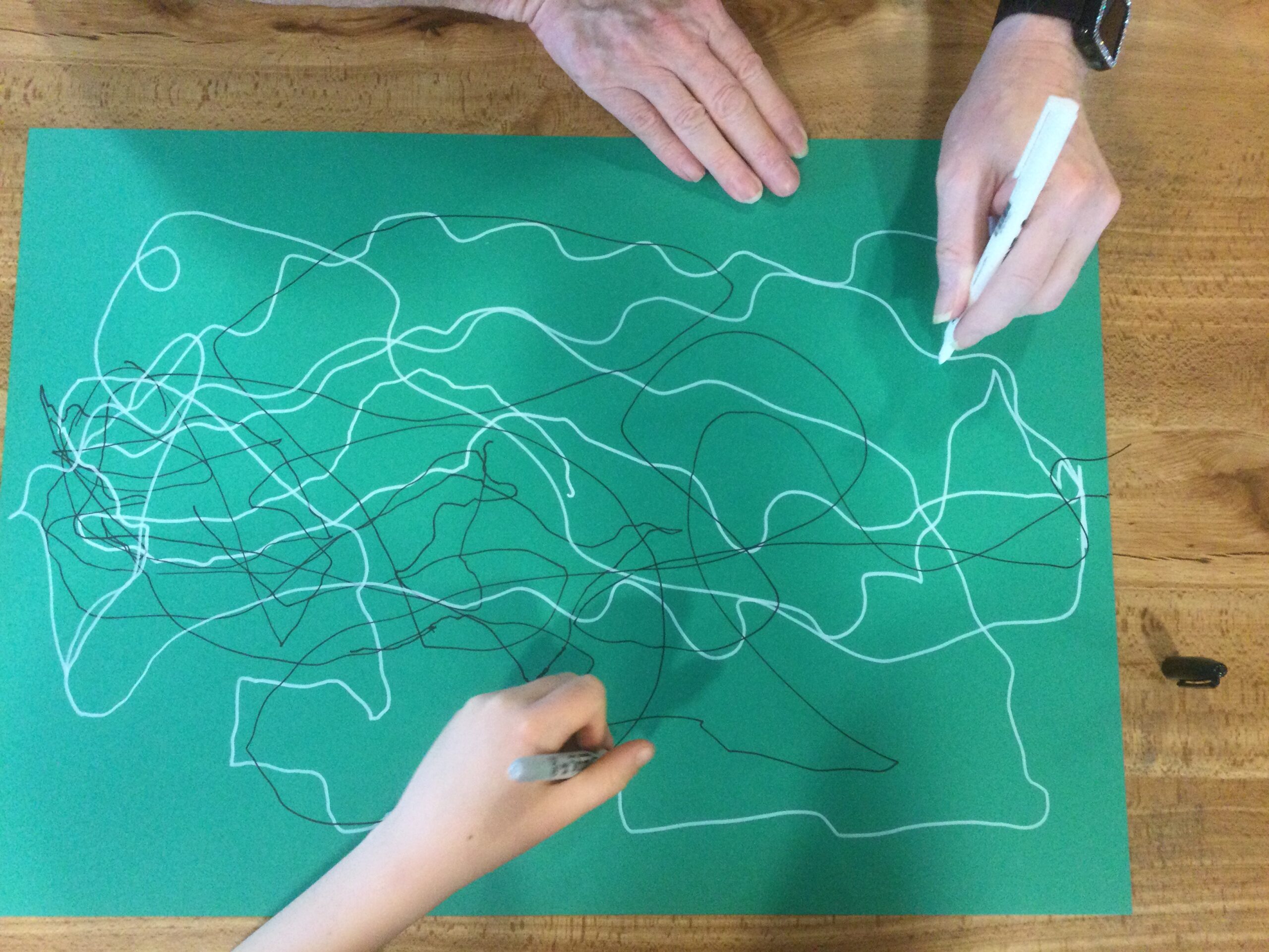 Overhead view of two pairs of hands drawing black and white lines on a green piece of paper