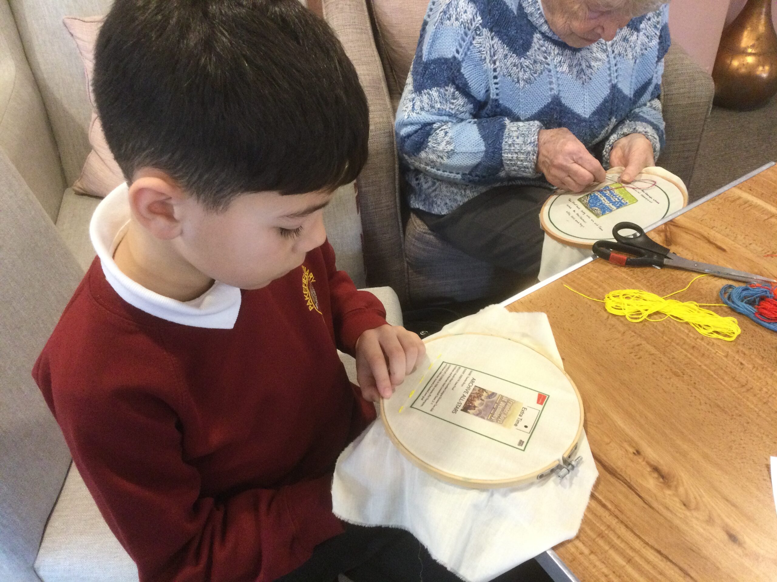 A child holding a printed piece of fabric in an embroidery hoop, he's sitting next to a woman stitching a picture