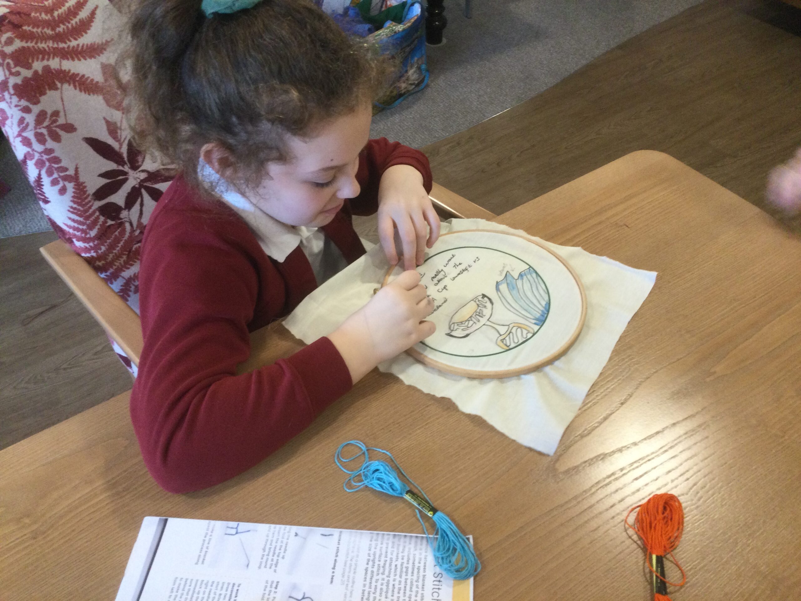 A school girl seated at a table, embroidering a piece of printed fabric