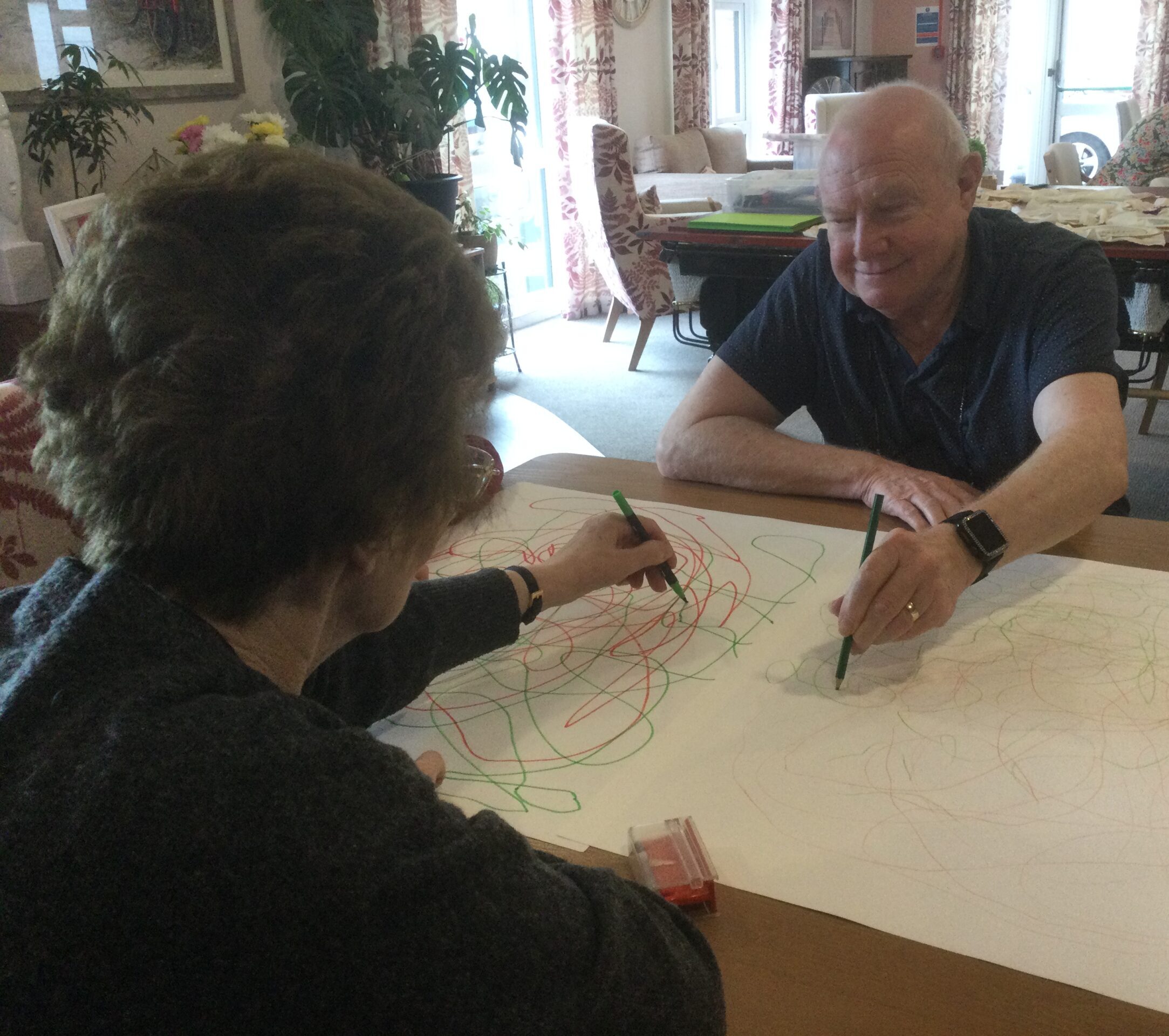 A gentleman smiles as he draws on a large piece of paper, seated opposite a woman, also drawing random lines