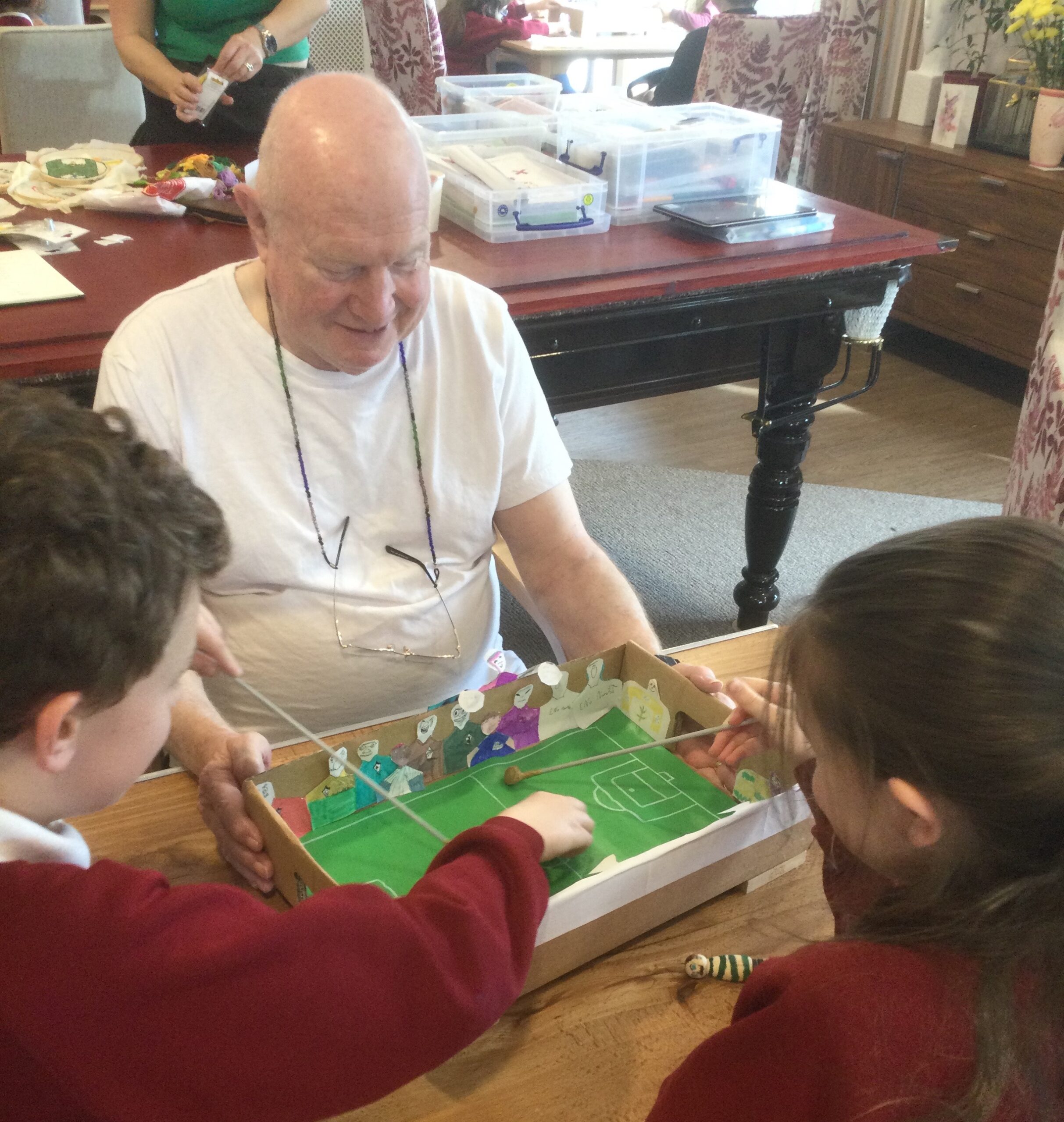 A gentleman in a white T-shirt watches on as two children play a table top football game