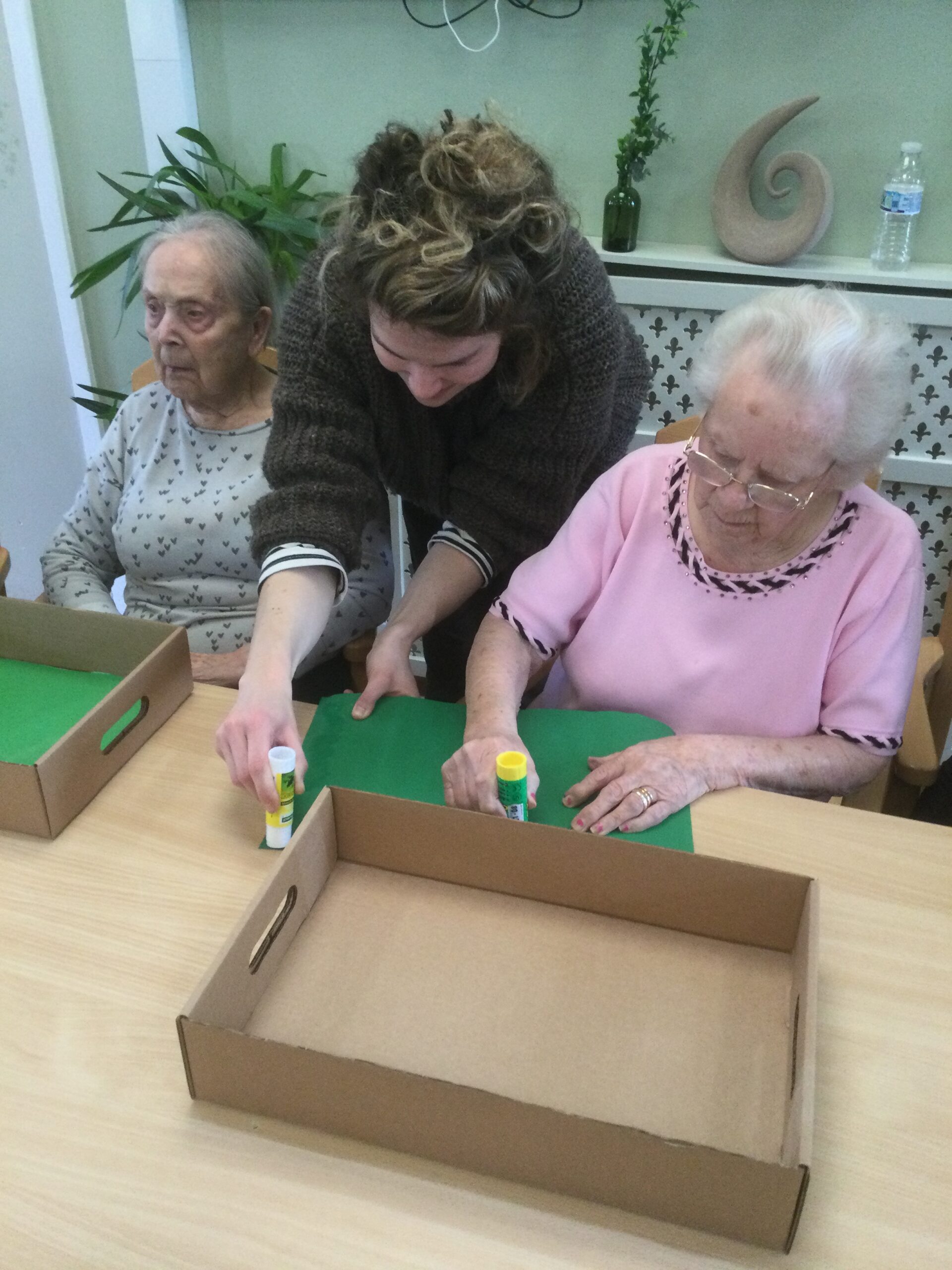 A woman leans between two ladies, applying glue to a piece of green material