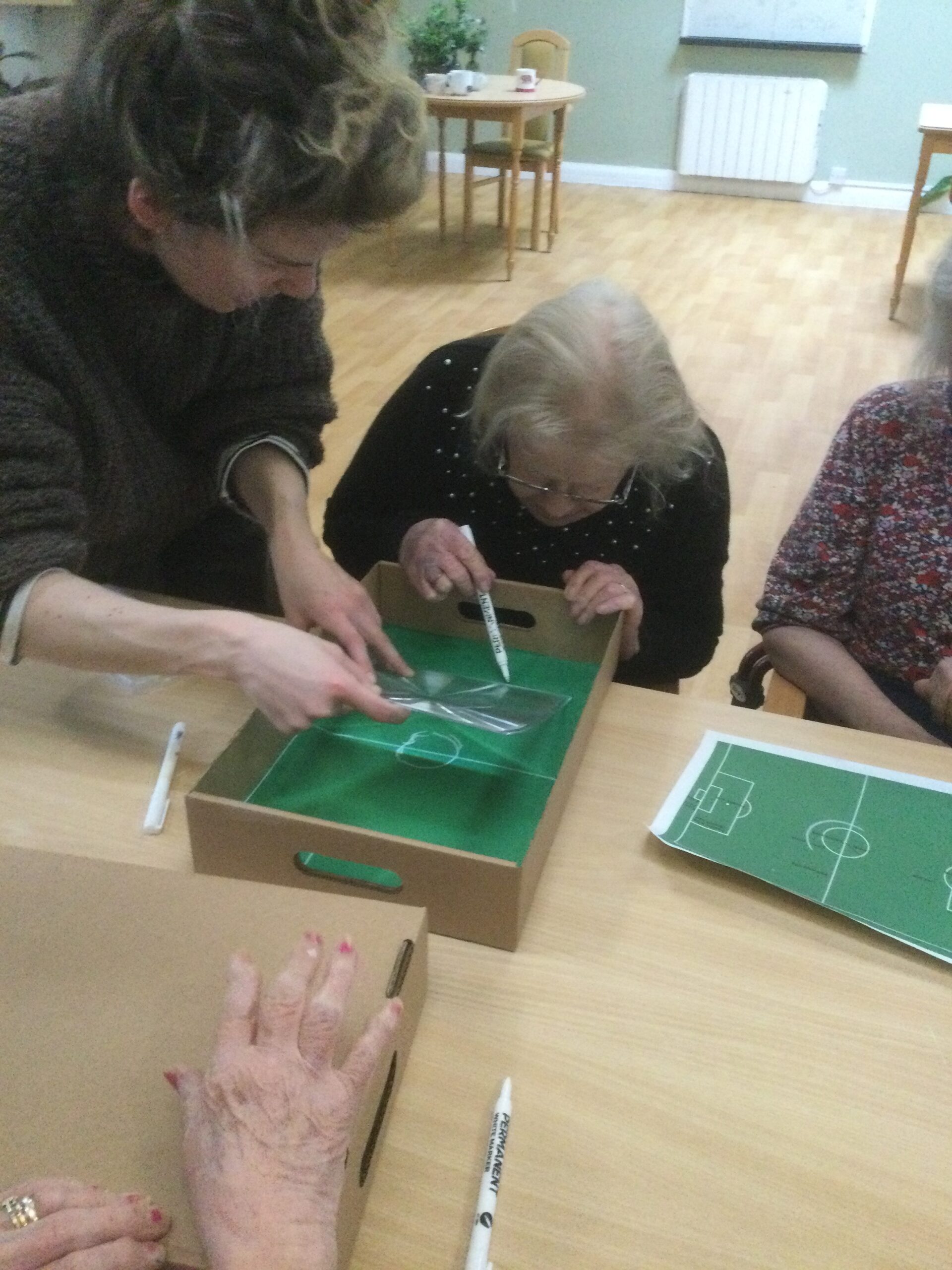 A woman bends over the box, drawing lines in white pen whilst another woman stands beside her, assisting