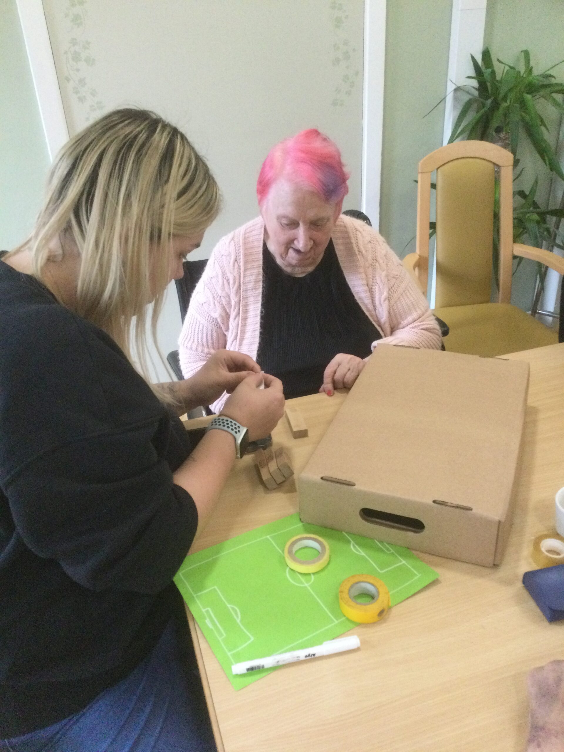 Two women seated at a table, with an inverted cardboard box in front of them applying something to the base