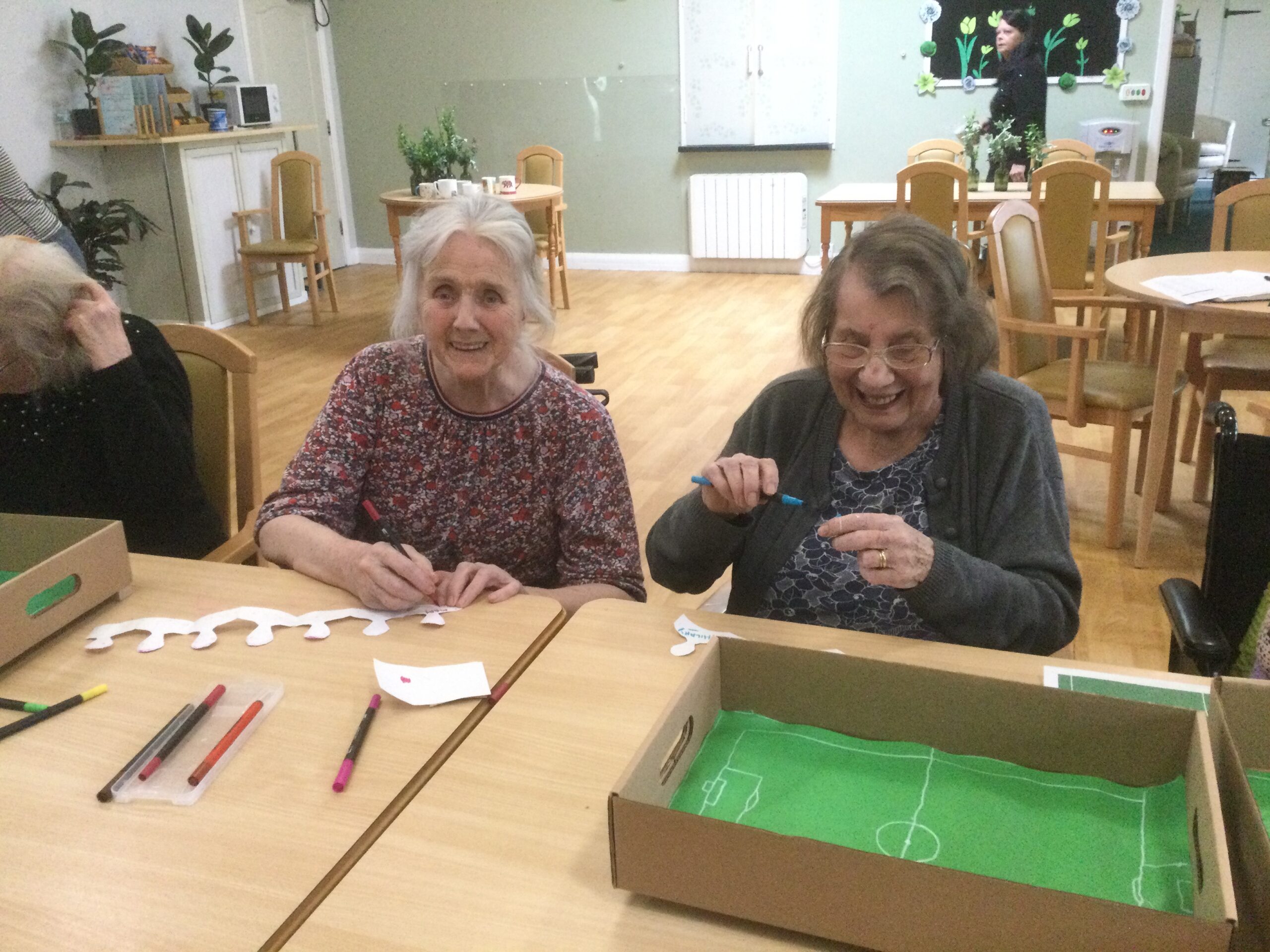 Two smiling women seated side by side, drawing a line of figures