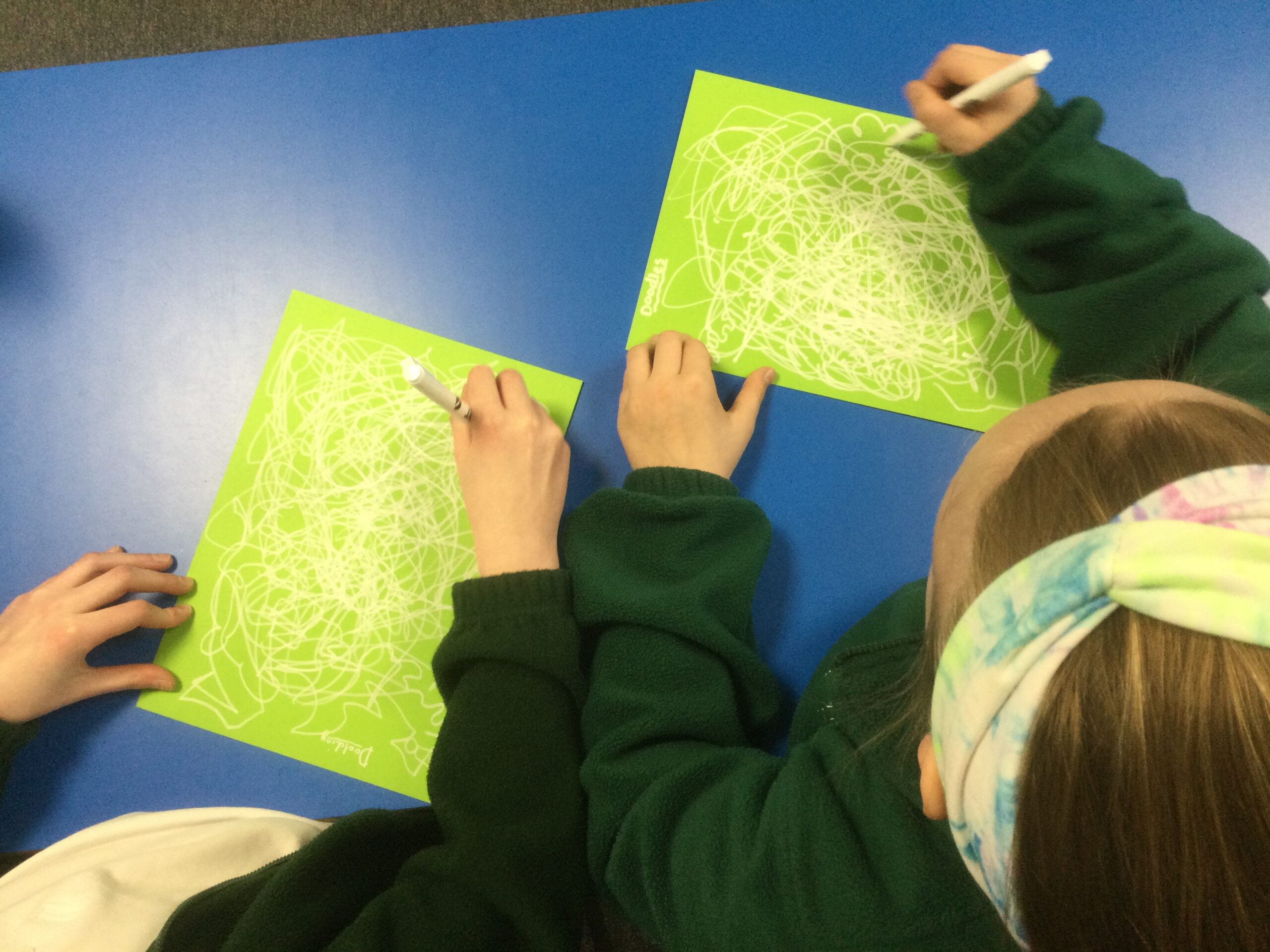 Overhead view of children drawing in white pen on a green background