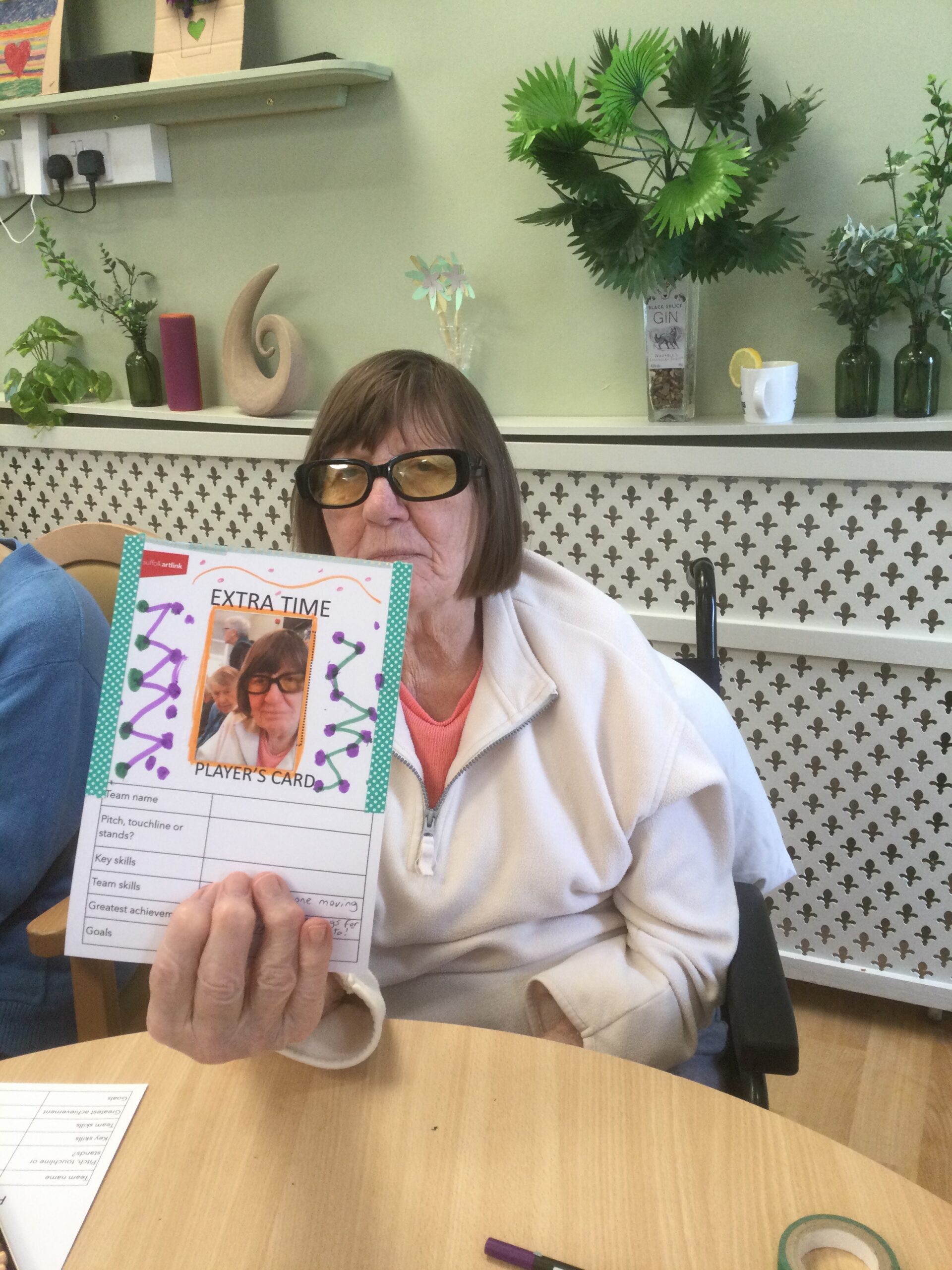 A woman seated at a table, holding up a hand-decorated card featuring an image of herself