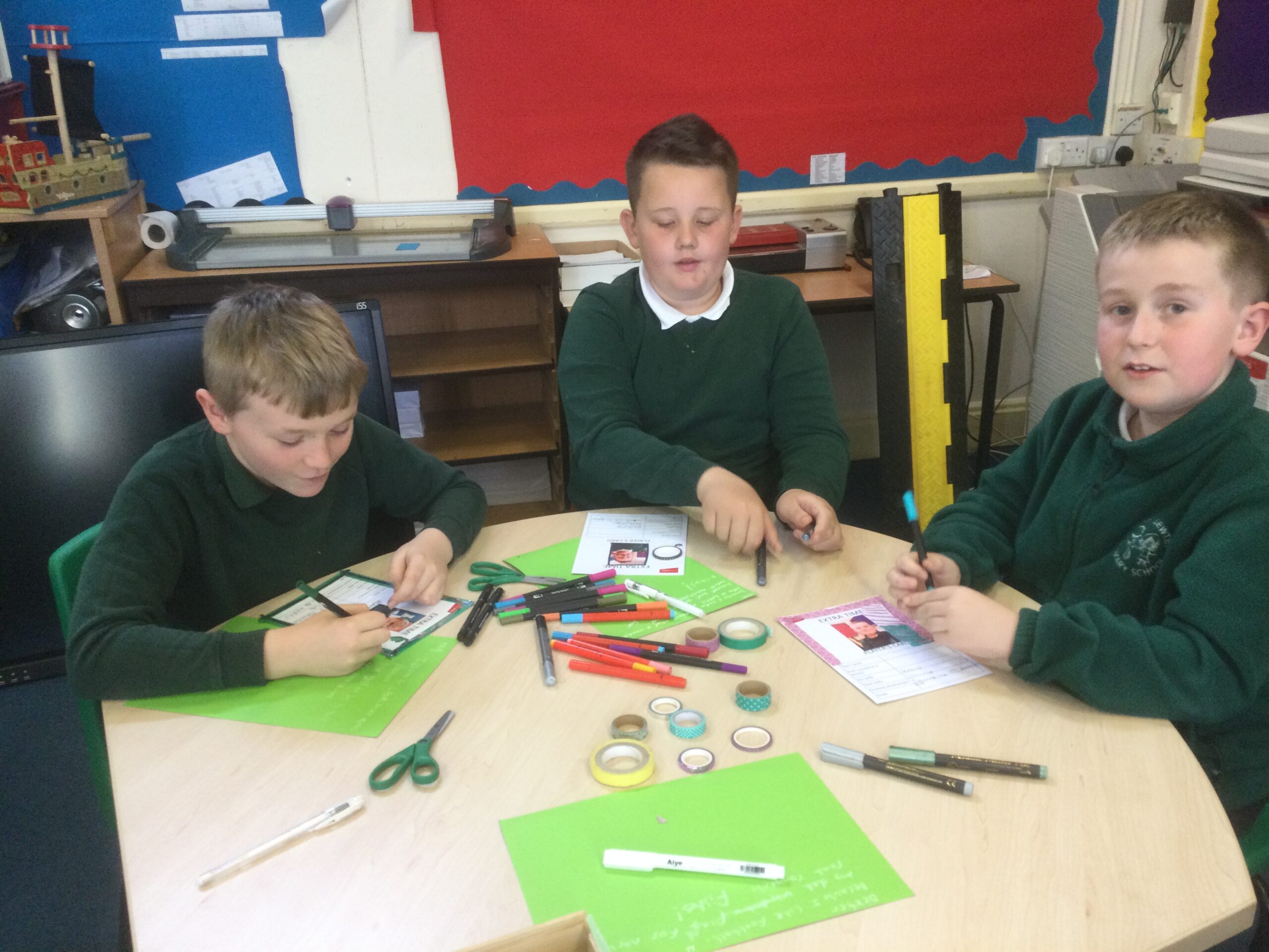 Three school boys in green jerseys decorating cards with coloured pens and tape