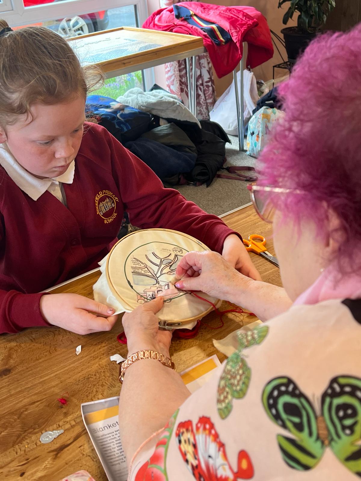 A woman stitches into a piece of fabric whilst a school girl opposite her holds the fabric taut